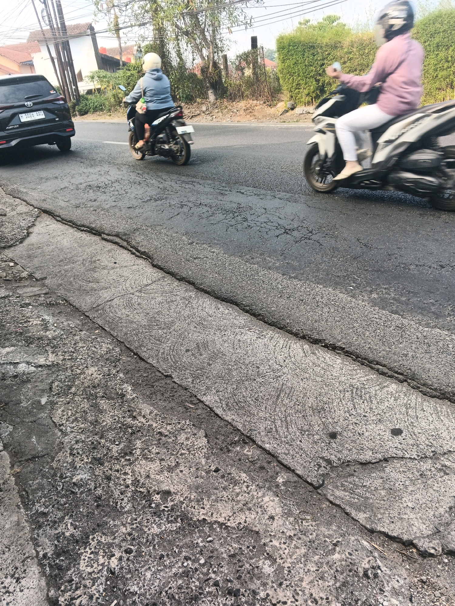 In Ungaran, Indonesia, a two-lane asphalt road is visible, with two motorcycles and one car traveling on it. The road has a rough texture with cracks and uneven patches. On the left side of the road, a black car with license plate H 1026 ADG is parked. To its right, a person wearing a blue hoodie and a white helmet is riding a motorcycle. Further down the road, a person in a pink shirt and white pants, also wearing a helmet, is riding a white motorcycle. The background features houses, trees, and a bamboo hedge. The scene appears to be a typical street in a residential area.