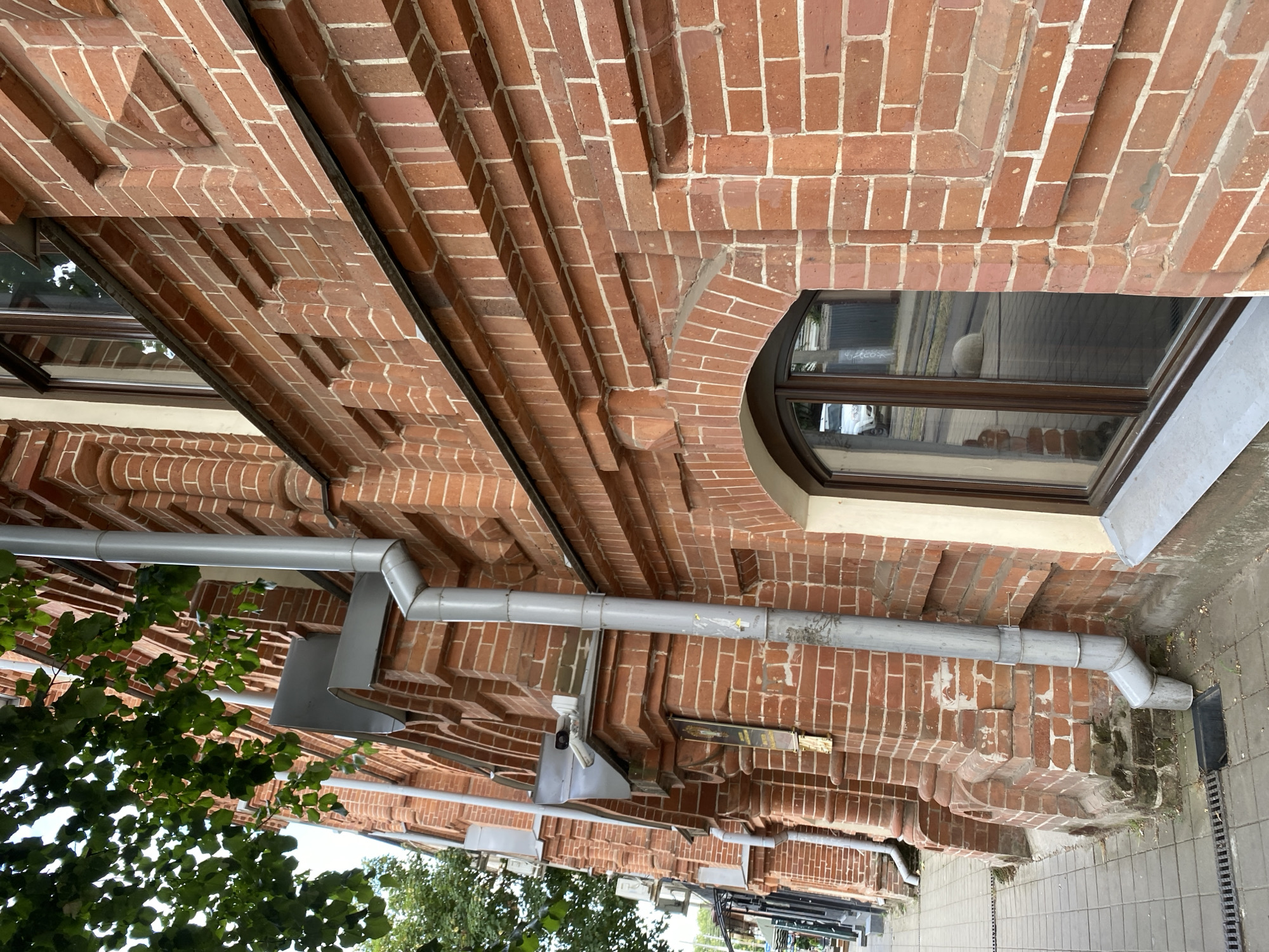 A low-angle, close-up, exterior shot shows the side of a red brick building, with a drainpipe running vertically down its facade. The building has an arched window with dark wood trim and the reflection of a white car and sidewalk is visible in the glass. Green leaves from a tree hang into the upper left of the frame, obscuring part of the building. The ground in front of the building is paved with square tiles and includes a drainage grate. The building appears to be located in a city, possibly Krasnodar, Russia, based on architectural style and typical urban elements.