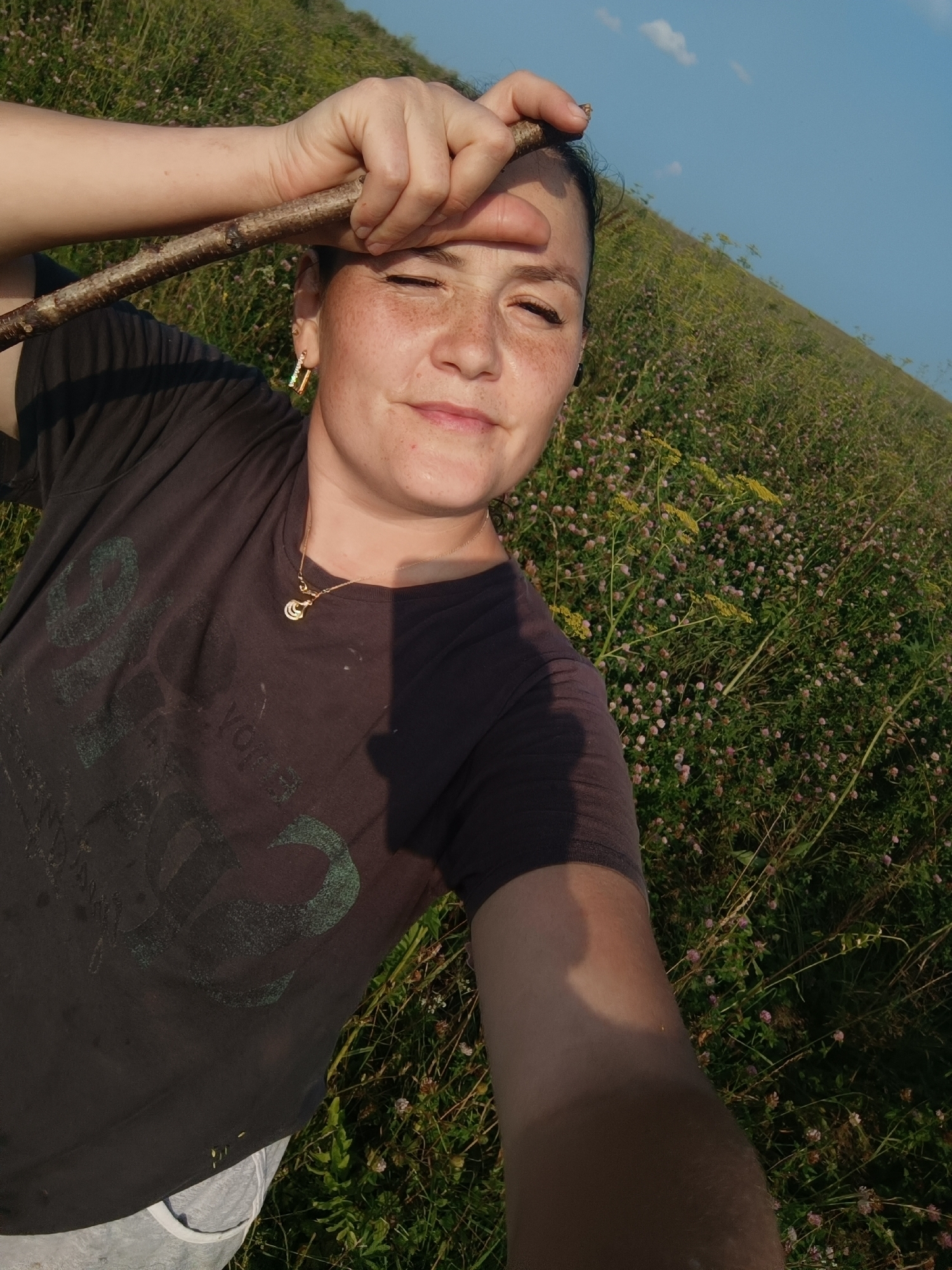 A woman is taking a selfie in a field of green grass and small pink flowers. She is wearing a dark t-shirt, grey shorts, and a gold necklace. A stick is held across her forehead. She has freckles on her face and is wearing an earring. The sun is shining, casting a shadow on her t-shirt, and the sky is blue with a few clouds. The scene appears to be outdoors, possibly in a rural area, with the sun suggesting it is daytime. The woman's expression is relaxed and content.