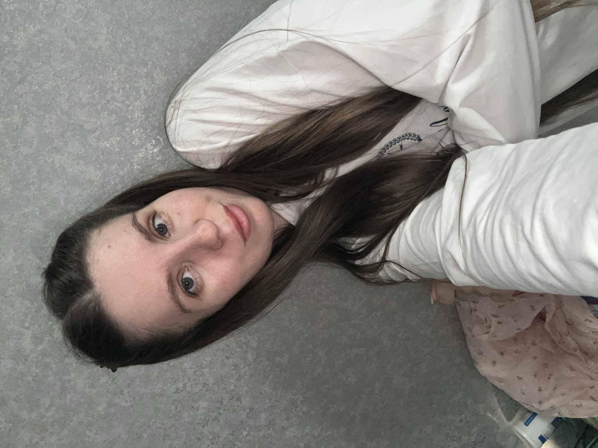 A young woman with long brown hair is lying on a gray surface, possibly a floor. She is partially covered with a white pillow or blanket, and her face is visible. She is looking up at the camera. A floral-patterned garment is also visible. There is no information available to determine if this image was taken in Klin, Russia.