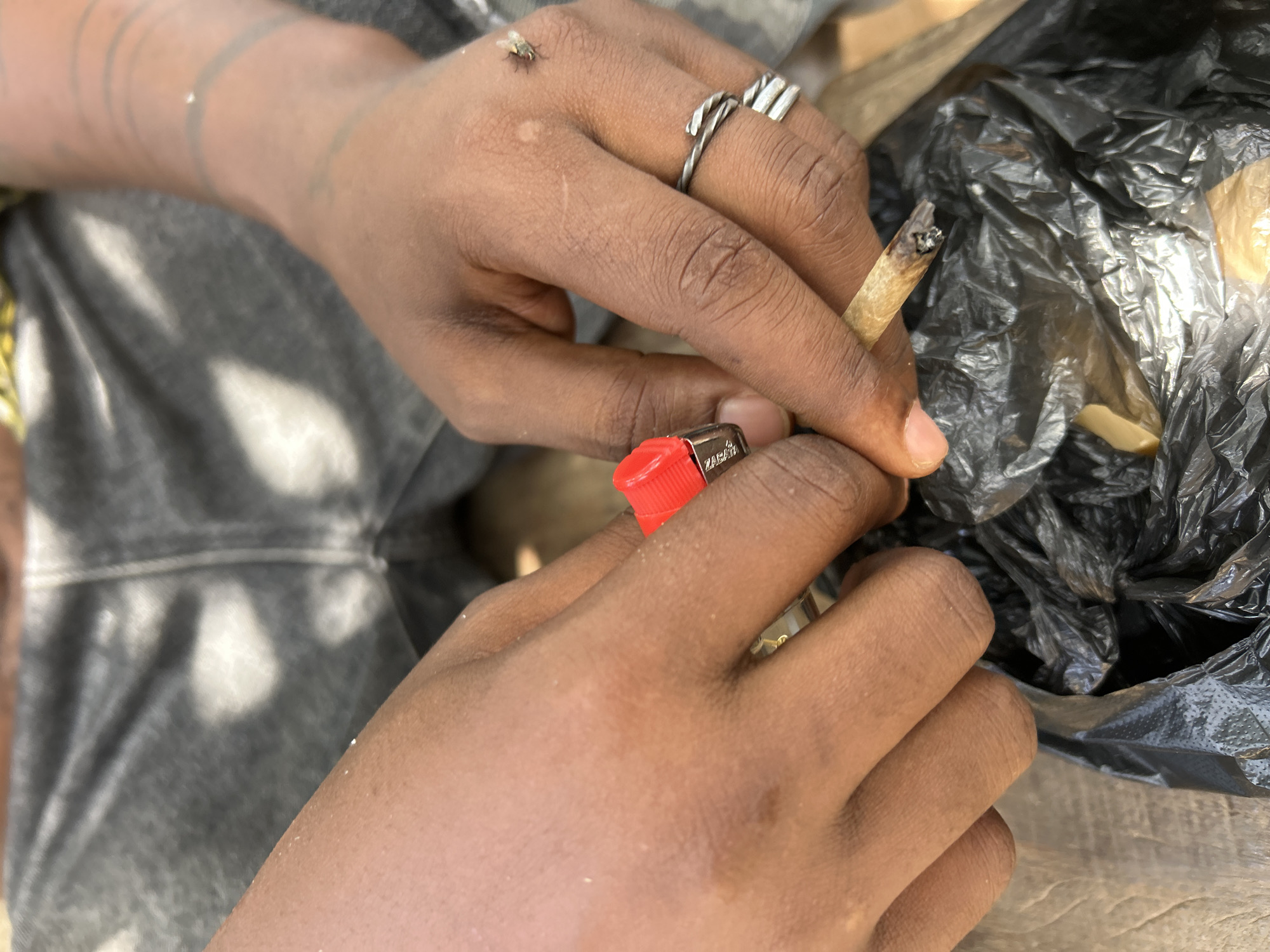 A person is seated outdoors, likely in Unknown, Unknown. They are holding a lit cigarette between their fingers and a red lighter. A black plastic bag is visible near their hand. A fly is perched on the back of their hand, which is adorned with two rings. They have tattoos on their wrist. The person is wearing grey pants.
