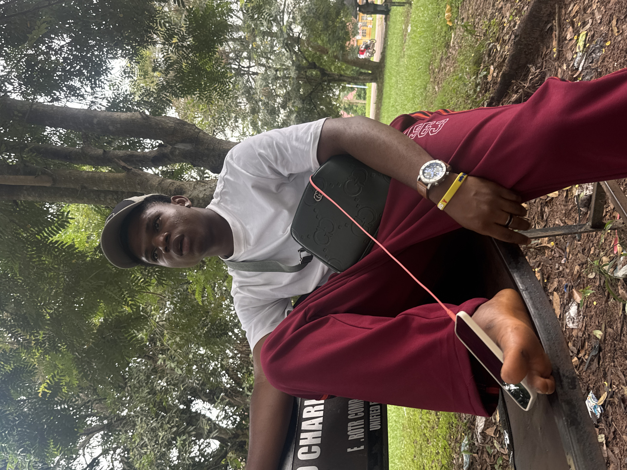 A young Black male is seated on a park bench in Ihiagwa, Nigeria. He is wearing a white t-shirt, maroon sweatpants, and a dark baseball cap. He is holding a dark-colored bag on his lap and has a smartphone resting on his foot. He is wearing a watch on his left wrist and a yellow bracelet. The bench has some text on it, partially obscured. The background shows trees, grass, and some distant buildings, suggesting an outdoor, possibly public, area.
