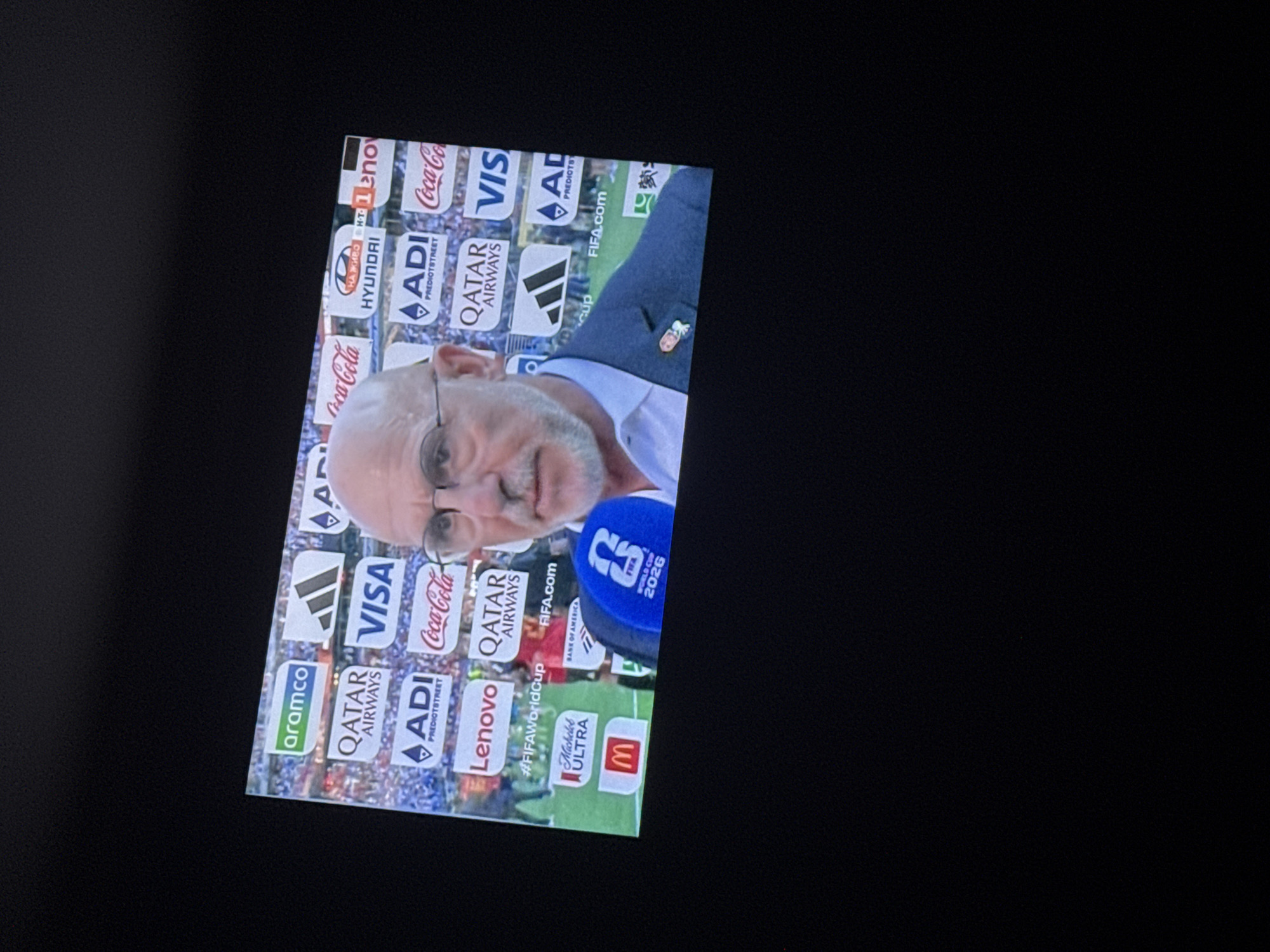 A man with glasses and a beard, wearing a suit jacket over a white shirt, is being interviewed with a microphone. He is standing in front of a backdrop of various sponsor logos, including Aramco, Qatar Airways, Visa, Coca-Cola, Hyundai, and FIFA. The backdrop appears to be a large screen or banner, set up in a stadium or event venue. The man is holding a microphone with a FIFA World Cup 2026 logo. The image is a medium shot, with the man's face and upper body filling most of the frame. The background is slightly out of focus, emphasizing the subject. The lighting is artificial, suggesting an indoor or evening event. There is no visible indication of the city of Krivodol, Bulgaria.