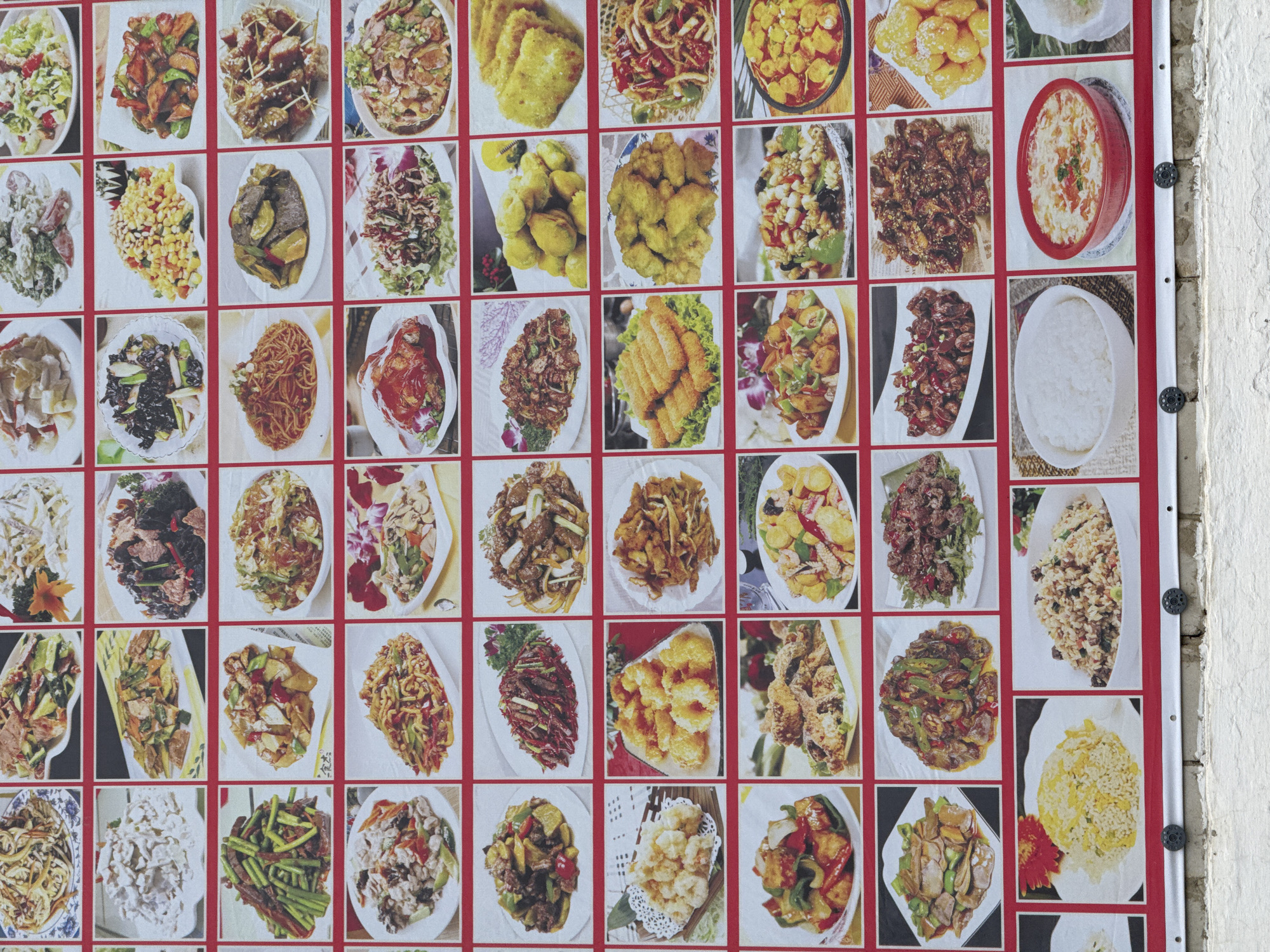 The image displays a vibrant, large-format menu board showcasing a variety of Chinese dishes. The board is divided into a grid of numerous individual photographs, each featuring a different culinary preparation. These photographs are arranged in rows and columns and are bordered by red frames. The dishes themselves appear to be well-lit and appetizing, with a diverse range of ingredients and cooking styles evident. Many of the dishes feature stir-fried vegetables, meats, and noodles, while others include fried items, rice dishes, and soups. The backdrop of the menu board is white, with a prominent red border. Below the menu board, a textured white brick wall is visible, with two black circular fasteners attached to the wall at the bottom of the menu board. The scene is brightly lit, suggesting an outdoor setting or a well-lit indoor space. The overall impression is that of a restaurant menu designed to attract customers with appealing visual representations of their offerings. The location is specified as Chuguyevka, Russia.