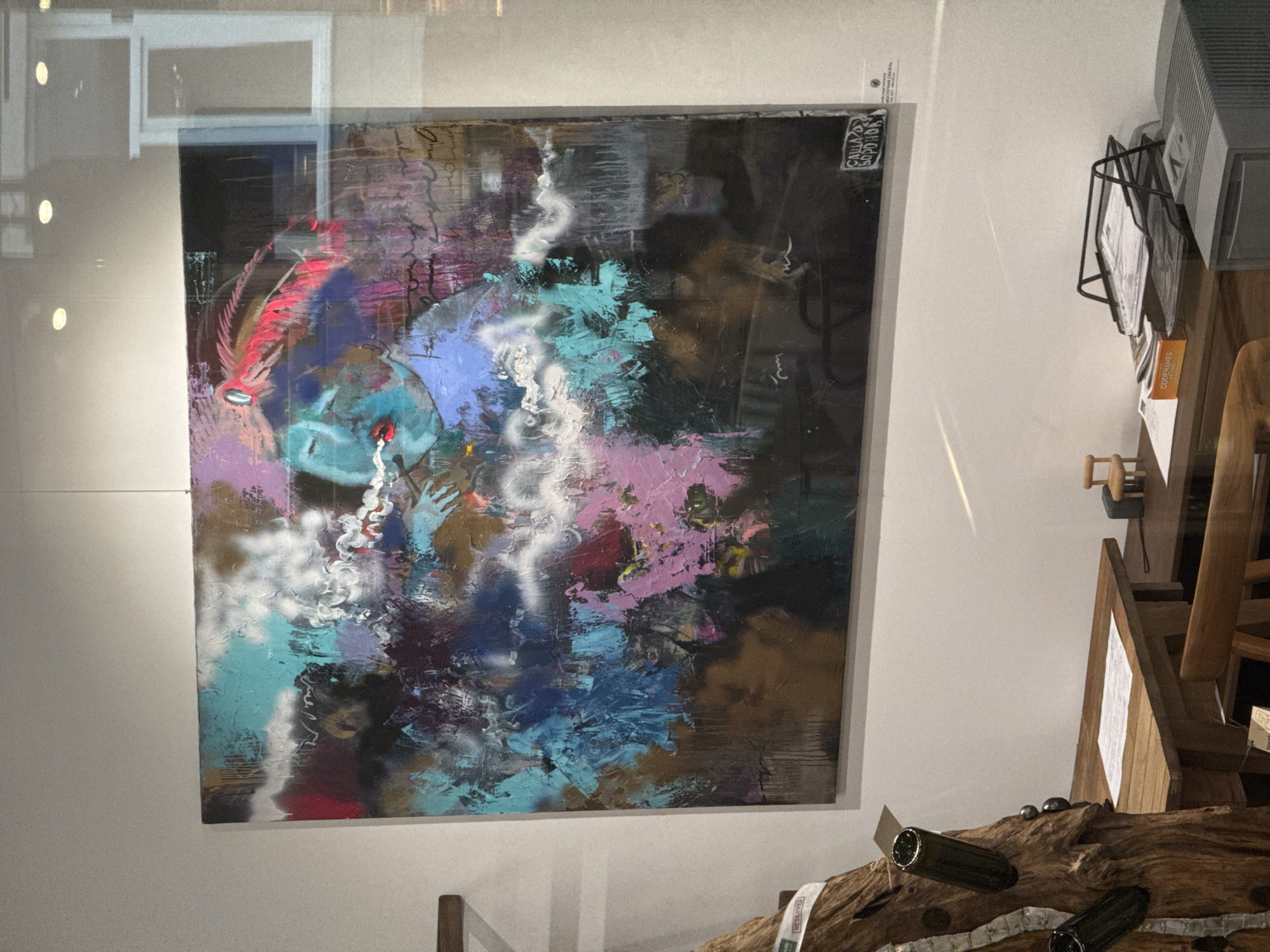 This image shows a painting with abstract brushstrokes in vibrant colors like pink, blue, and turquoise, against a dark background. The painting appears to be framed and hung on a white wall. There are reflections on the surface of the painting, suggesting it might be behind glass, possibly in a display window or a gallery. To the right of the painting, there are wooden shelves with various objects, including a metal magazine rack filled with papers and a computer monitor. The lighting suggests an indoor setting, possibly during the day or early evening. On the top right corner of the painting, there is a small white label with text, which appears to be a signature or artist's mark. The overall scene evokes an artistic and perhaps commercial environment.