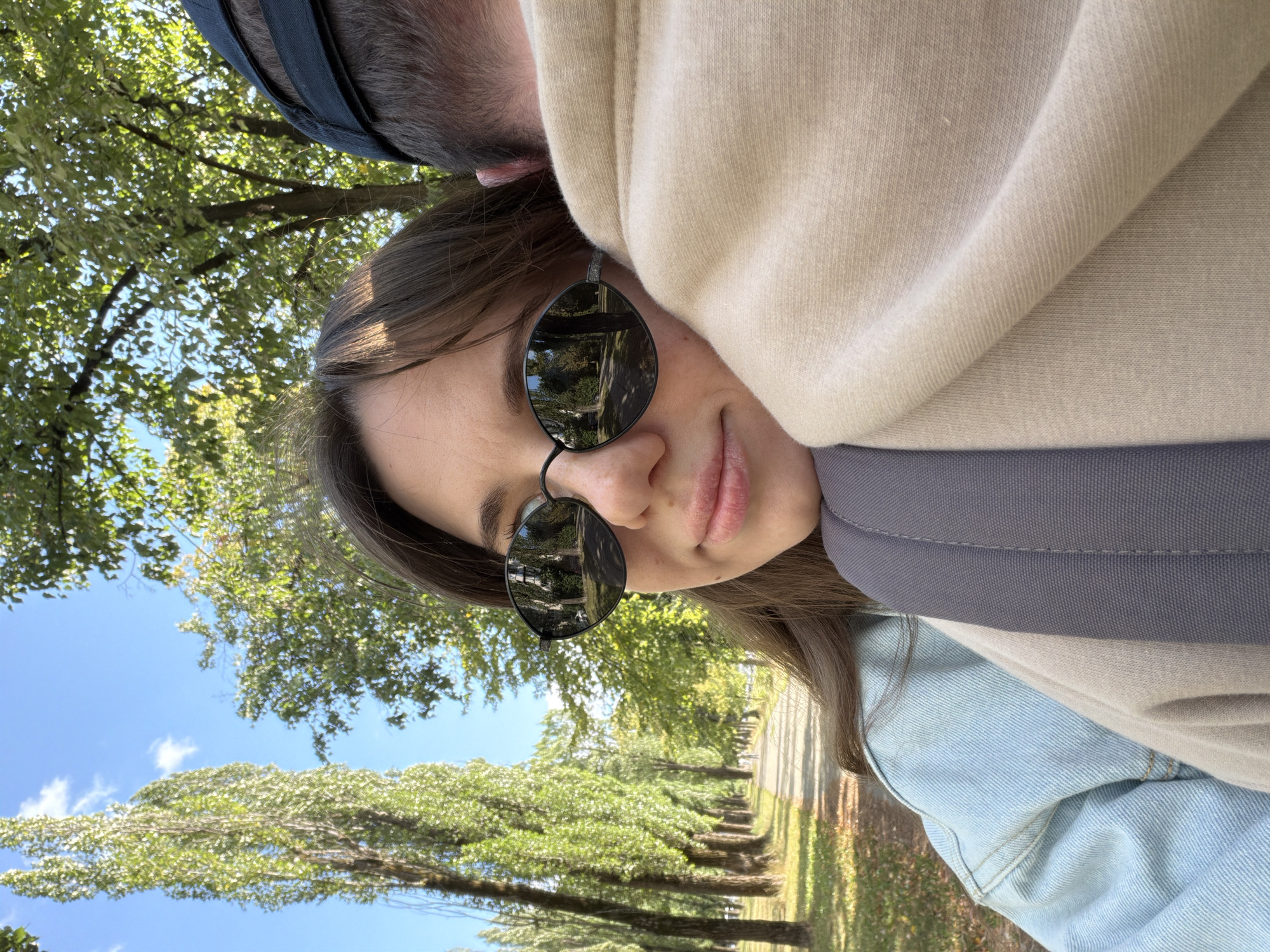 This is a close-up, outdoor, selfie-style photograph taken on a sunny day in Kyiv, Ukraine. The foreground features a young woman with brown hair, wearing sunglasses and a light blue denim jacket, her head resting on the shoulder of an unseen person who is wearing a beige hoodie and a dark baseball cap. The background depicts a tree-lined path, likely in a park or boulevard, with tall, slender trees flanking the walkway. The clear blue sky is visible through the green foliage. The reflection in the woman's sunglasses shows the same tree-lined path, suggesting the photo was taken while walking. The overall impression is a casual, intimate moment shared between two people outdoors.