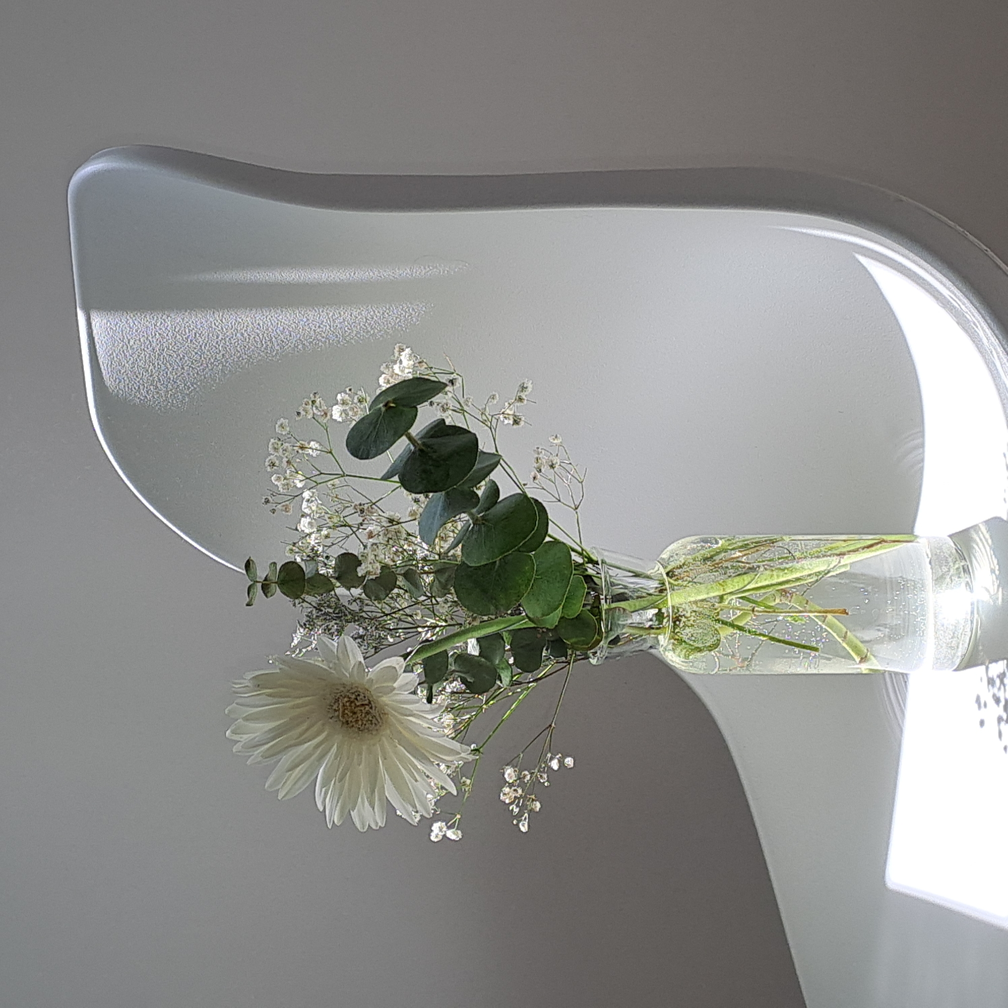 A close-up shot shows a clear glass vase with a bouquet of white flowers and eucalyptus branches. The vase is filled with water and sits on a surface that reflects light. The bouquet features a large white gerbera daisy as the focal point, surrounded by delicate white baby's breath and several rounded green eucalyptus leaves. The arrangement is illuminated by natural light, casting shadows on the plain, light gray background. The right side of the image is dominated by a white curved object, possibly a chair or a piece of furniture, which is partially in shadow and partially illuminated, creating a textured surface with speckled light. The overall scene is minimalist and serene, with a strong emphasis on light and shadow play. No people are present in the image. The location is Unknown, Unknown.