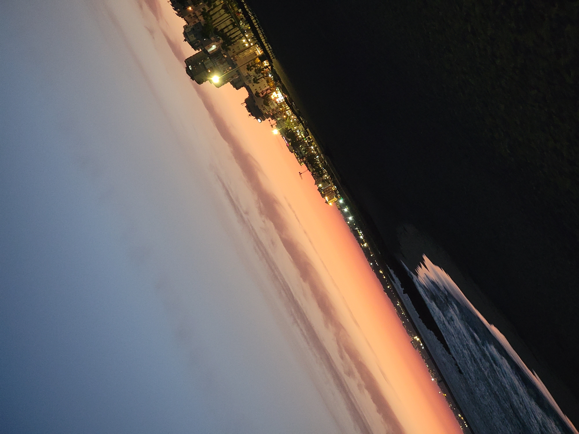 The image captures a twilight view of the Quarteira coastline in Portugal. The sky is a gradient of deep blue at the top transitioning to a soft orange and pink horizon, indicating sunset or sunrise. Wispy clouds are visible in the sky.
Along the coast, buildings are illuminated, creating a glow against the darkening sky. These buildings appear to be hotels or apartment complexes, with rows of lights suggesting occupied rooms. Palm trees are interspersed among the structures, a common feature of the Algarve region.
The foreground of the image consists of a sandy beach and the ocean. The water is dark, reflecting the dim light of the sky. Gentle waves are visible, breaking onto the shore. A breakwater or rocky structure extends into the sea, partially submerged by the water. The overall scene depicts a serene coastal town at dusk.