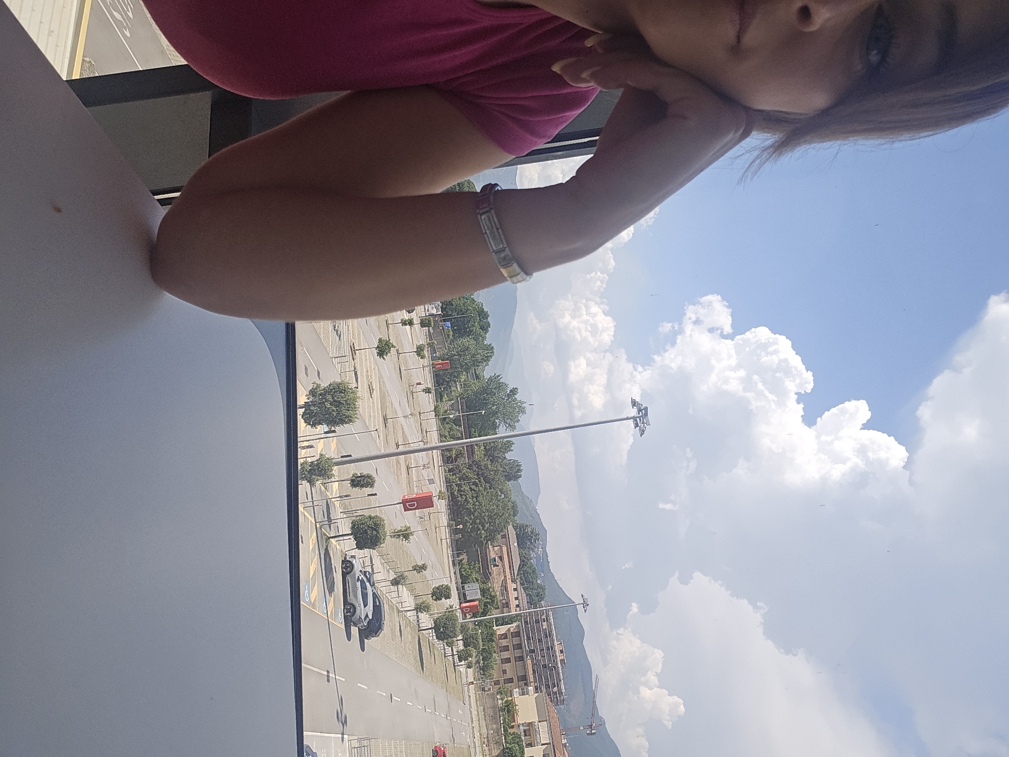 A woman wearing a bright pink t-shirt is captured in a selfie, her face partially visible as she rests her chin on her hand. A silver bracelet adorns her wrist. Behind her, a vast, nearly empty parking lot stretches out, lined with young trees and illuminated by tall lampposts. In the distance, a cluster of buildings and lush green hills rise under a bright, cloudy sky. The scene is identified as being in Baronissi, Italy, suggesting the parking lot may be adjacent to a commercial or public area within the city.