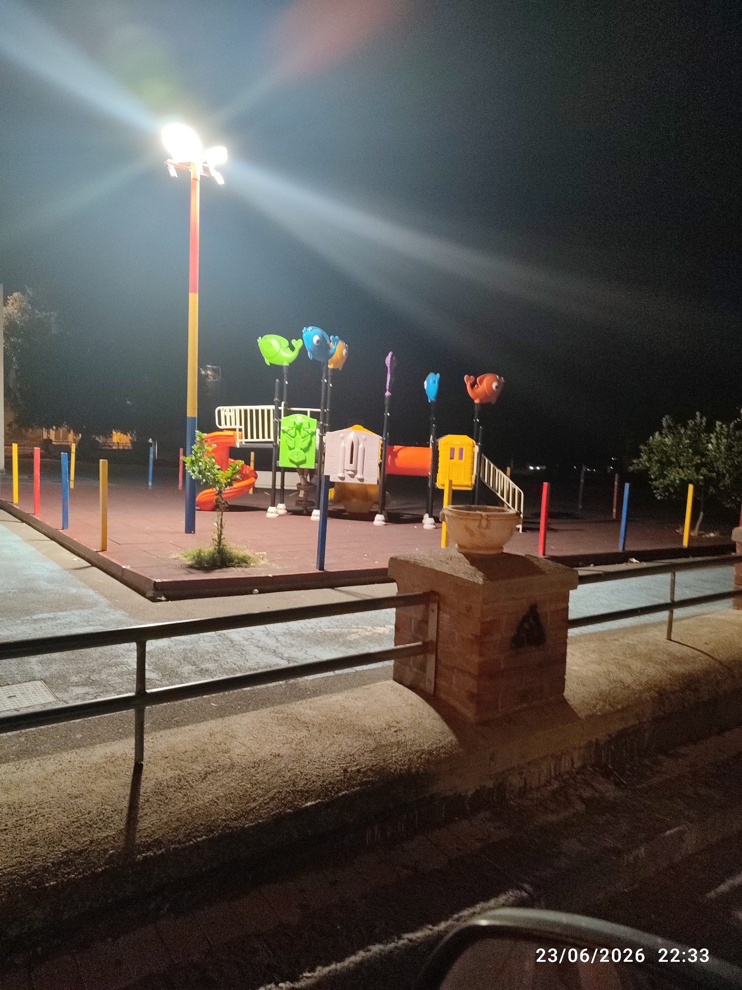This media file depicts a brightly lit playground at night, likely in Scordia, Italy, as indicated by the timestamp "23/06/2026 22:33". The scene is illuminated by a tall, multi-colored pole light that casts strong beams and lens flares across the dark sky. The playground equipment is colorful, featuring what appear to be stylized animal-shaped swings or decorations in shades of green, blue, and orange, along with slides and climbing structures. The ground of the playground is covered with red rubber matting.
In the foreground, a low brick structure with a circular planter on top is visible, along with a metal railing. The background shows more of the playground with colorful poles marking its perimeter, and a glimpse of trees and distant lights, suggesting an urban or suburban setting. The overall atmosphere is one of a quiet, well-maintained public space after hours. There is no activity or people visible in the frame, giving the scene a sense of stillness. The image is taken at night, with the artificial light being the primary source of illumination. The weather appears clear, as indicated by the sharpness of the light beams.