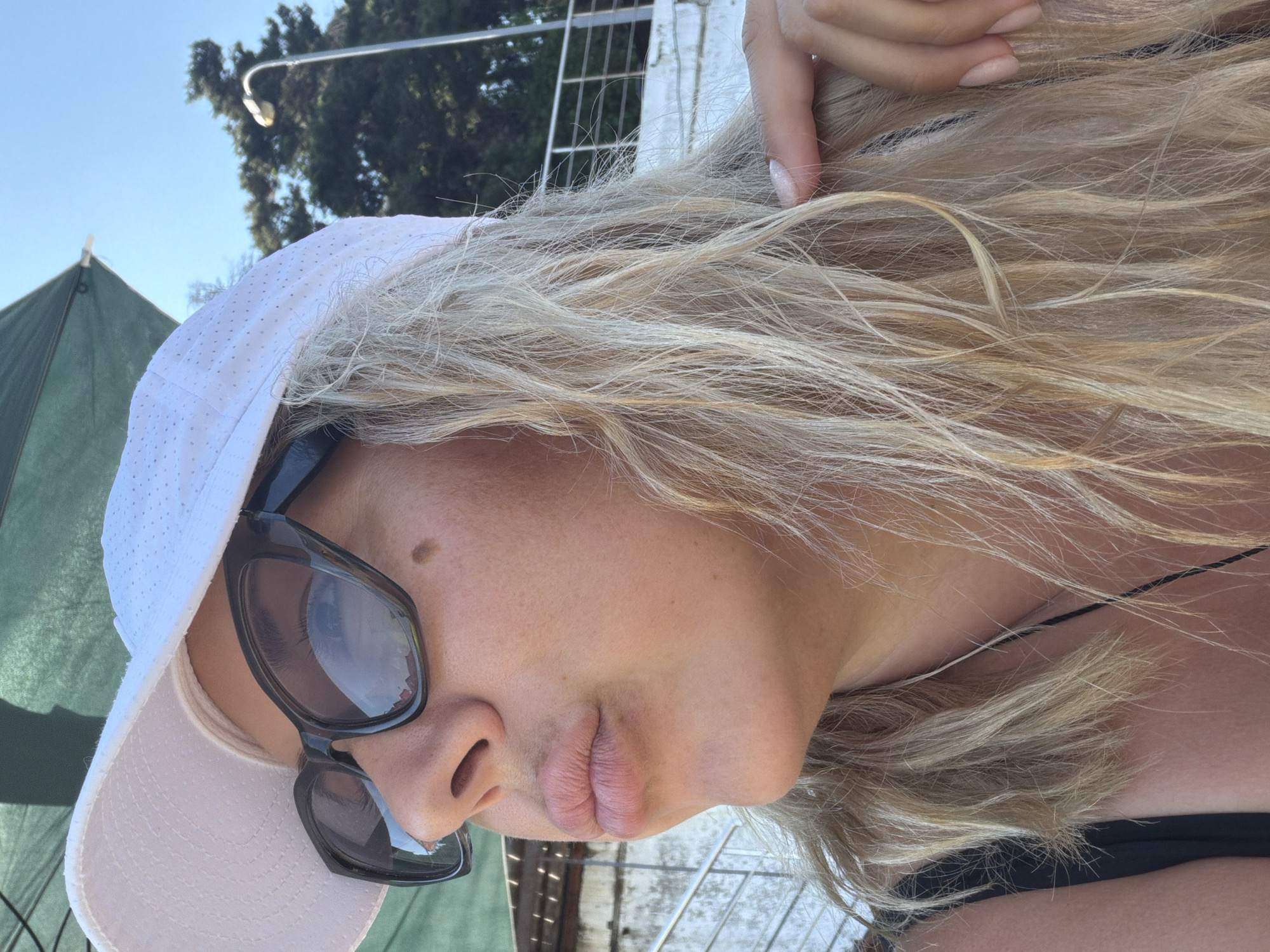 The image shows a close-up, sideways selfie of a woman with blonde hair. She is wearing a white baseball cap and dark sunglasses. Her hair is partially covering her face and shoulders. A hand with painted nails is visible touching her hair near the top of her head. She appears to be outdoors, with a blue sky and some trees visible in the background. A green tent-like structure is to the left of the frame. The lighting suggests it is daytime, likely sunny. The woman's facial features are partially obscured by her sunglasses and hair, but her lips are visible. The overall impression is casual and summery. There is no visible text in the image.