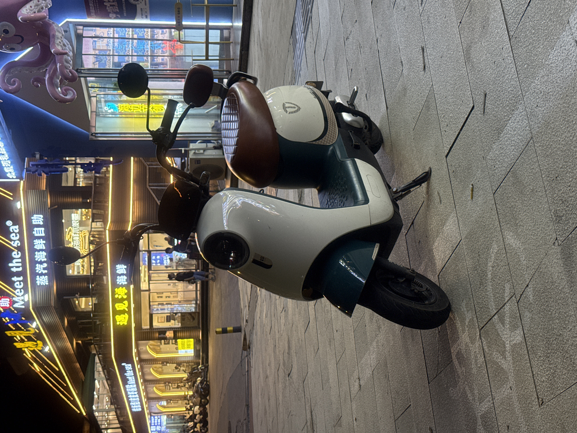 The image captures a street scene at night in Qinhuangdao, China. The focal point is a white and teal electric scooter parked on a paved sidewalk. Behind the scooter, a brightly lit building facade features signage in Chinese characters, including "Meet the sea" and "蒸汽海鲜自助" (Steam Seafood Buffet). The building has multiple levels and illuminated windows. To the right, a blue structure with a pink octopus decoration is visible, along with more illuminated storefronts. Several other motorcycles are parked further down the street to the left. The ground is made of large, light-colored stone tiles. Two individuals are dimly visible walking inside one of the buildings. The overall atmosphere is that of a commercial area at night, with neon lights and storefronts creating a vibrant ambiance.