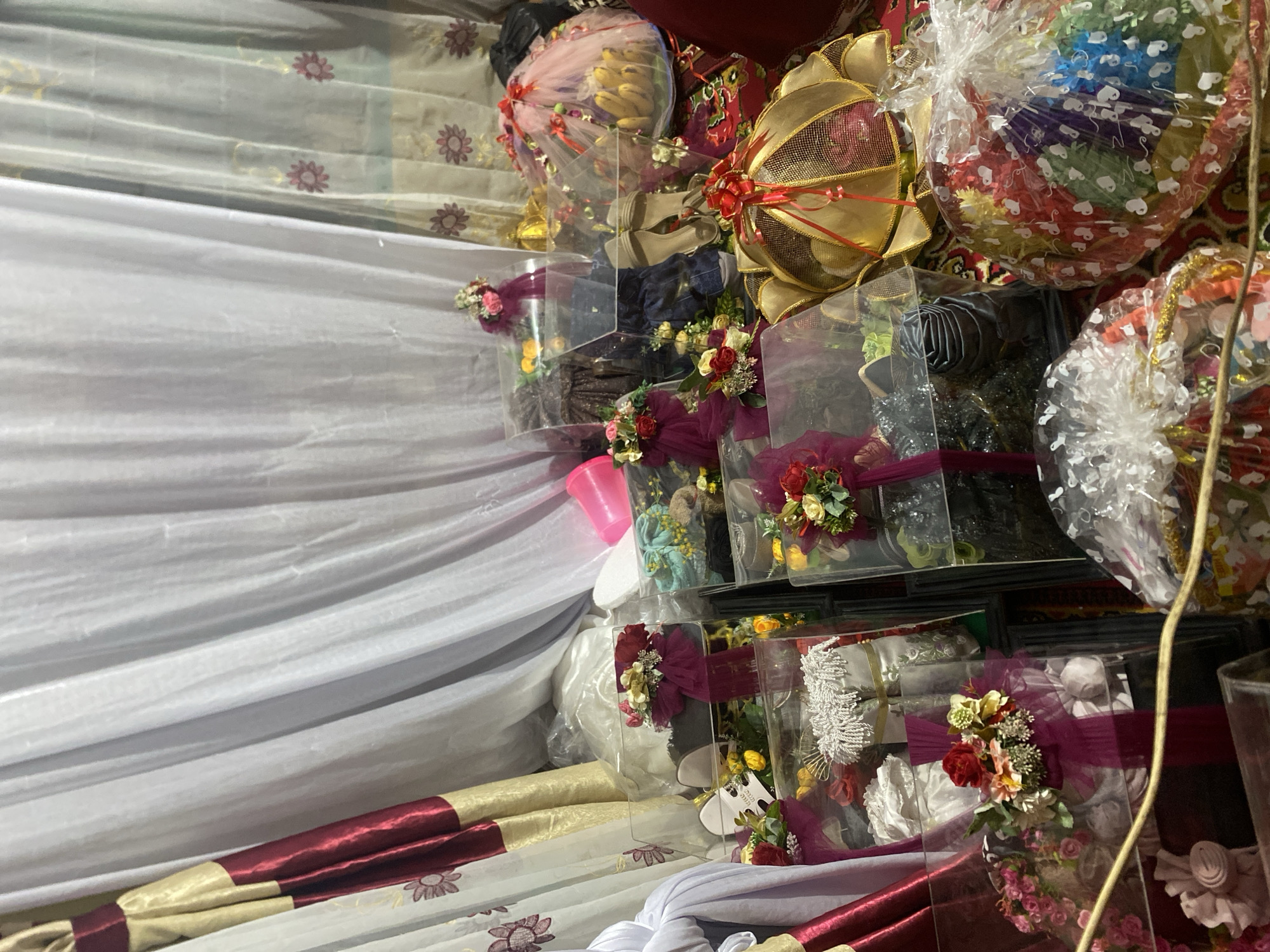The image depicts a collection of gifts, possibly wedding gifts, arranged on what appears to be a white and patterned fabric surface. The gifts are packaged in clear plastic containers, adorned with ribbons and floral decorations. Items visible inside the containers include clothing, shoes, and food items like bananas. The location is likely Seuneuboklhong, Indonesia, based on the prompt's request.
