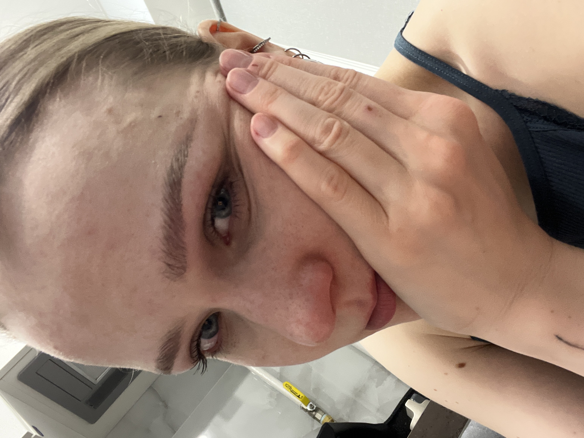 A close-up, slightly angled view of a person's face and part of their upper body. The person is fair-skinned with light brown hair and blue eyes. Their right hand is covering part of their face, specifically the forehead and cheek. The fingernails are short and appear to be painted with a neutral color. The person is wearing a dark-colored, thin-strapped top with lace trim. The background is predominantly white, with a glimpse of a utility pipe with yellow markings and a black object in the lower center. The surroundings appear to be indoors, possibly a bathroom setting. The image does not provide information to determine location in Khimki, Russia.