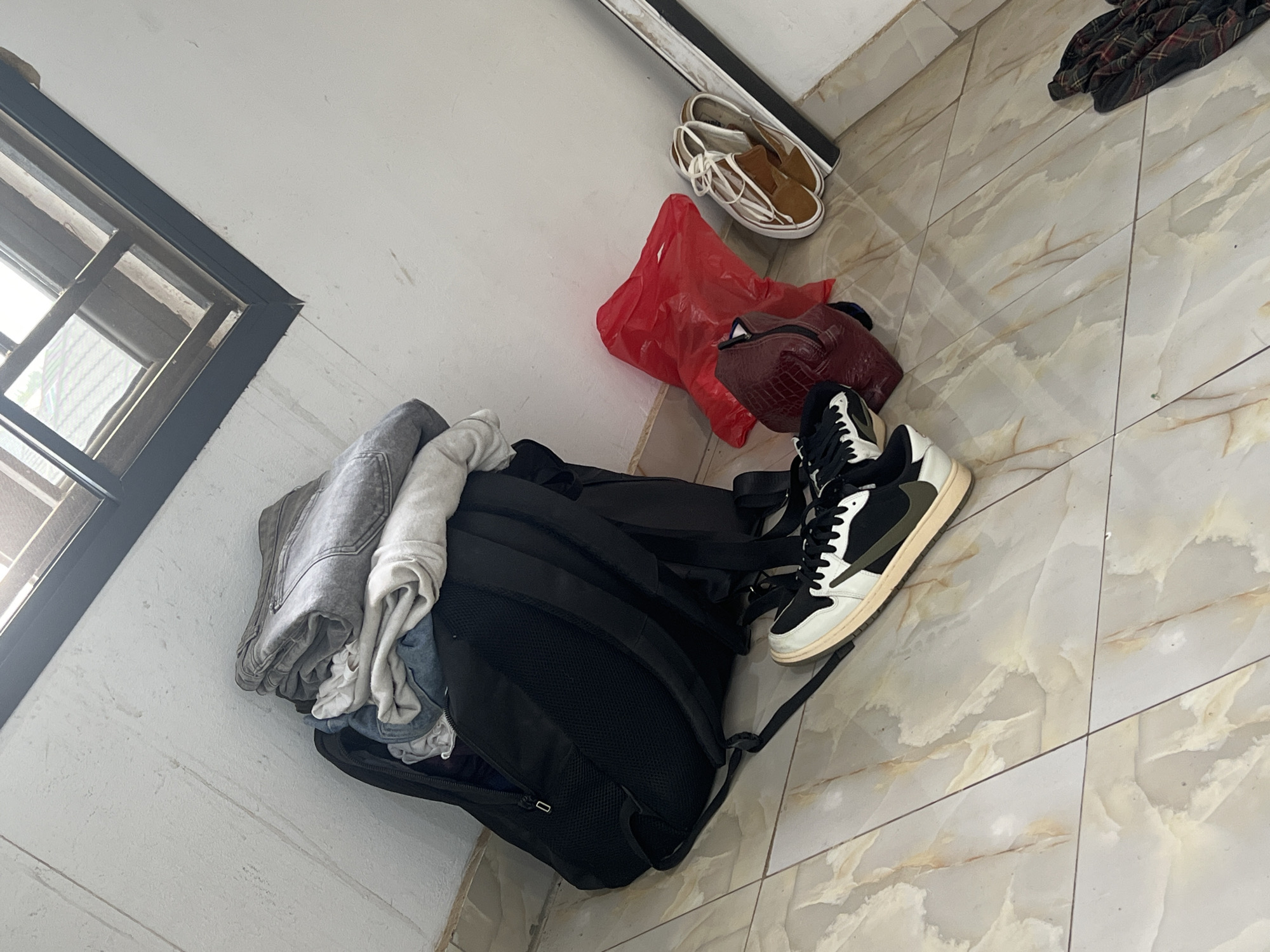 This image captures an indoor scene, likely a room in Abuja, Nigeria, from a high-angle perspective, showing various personal items arranged on a light-colored tiled floor.
The central subject is a black backpack, appearing partially open or having clothes placed on top. Rolled or folded items of clothing, including what looks like grey denim jeans and several light-colored t-shirts or fabric pieces, are visible on and around the backpack.
To the right of the backpack, there's a pair of black, white, and olive-green high-top sneakers. Further to the right, a maroon or burgundy handbag with a textured, possibly crocodile-skin-like pattern, is present. Above this, a crumpled red plastic bag is visible. Near the top right corner, a pair of light brown slip-on shoes with white laces rests against the wall or baseboard. A dark plaid fabric, possibly a shirt, is also partially visible in the far upper right.
The floor is covered with light-colored square tiles featuring a marbled pattern in shades of cream, beige, and light brown. The walls are plain white or off-white. In the upper left quadrant, a window with dark framing and multiple panes allows natural daylight into the room, suggesting it is daytime.
No people are visible in the image, and there is no discernible activity beyond the static arrangement of objects, possibly implying preparation for travel or unpacking. No text is visible in the frame.