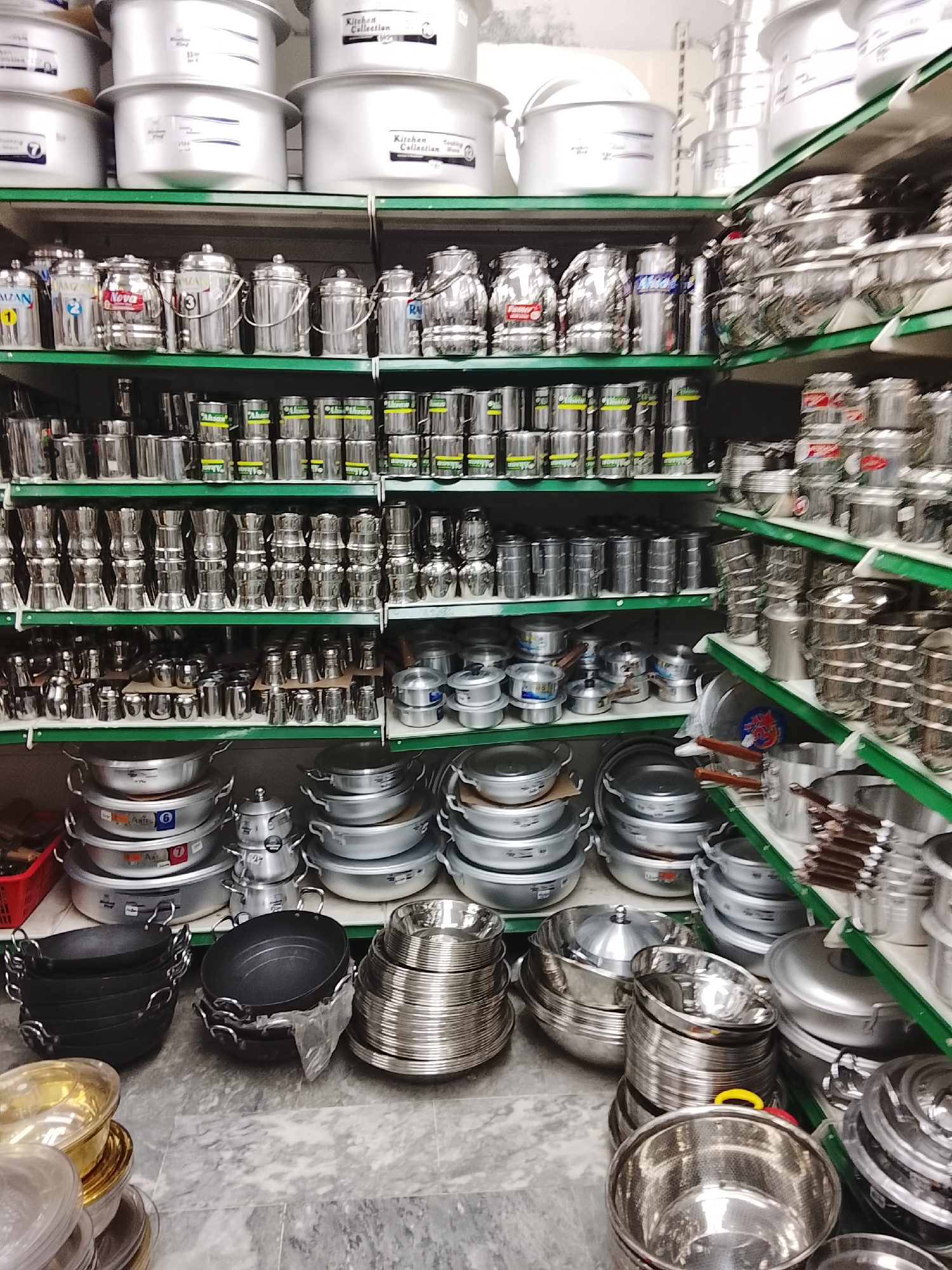 The image depicts the interior of a shop, likely in Gulgasht Colony, Pakistan, based on the query. The focus is on shelves heavily stocked with various types of metal cookware.
**Objects:**
* Multiple shelves holding stacks of aluminum and stainless steel pots, pans, and other cooking utensils.
* A variety of cookware types are visible, including large stockpots, smaller saucepans, bowls, and frying pans (some non-stick).
* Some items have labels or stickers on them.
* Shelving units with green supports.
* A tiled or marble-like floor.
**Environment:**
* The scene appears to be within a retail establishment, presumably a kitchenware or general goods store.
* The lighting is sufficient for viewing the merchandise, although perhaps somewhat generic.
No people are visible in the frame.