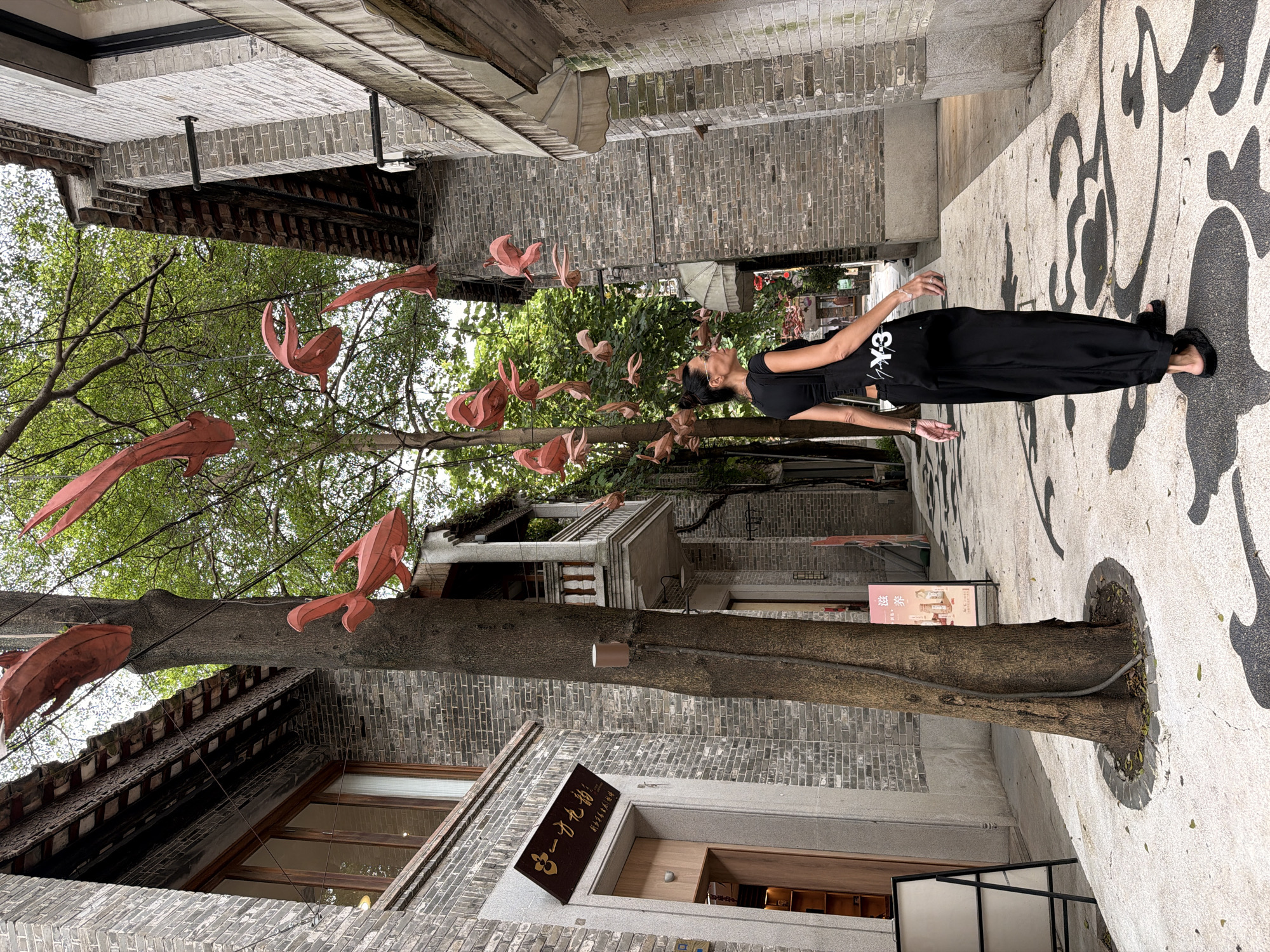 A woman walks down a pedestrian alley in Chancheng District, China, with a tree adorned with red fish-shaped lanterns in the foreground. The buildings on either side of the alley are constructed of grey brick with traditional tiled roofs. The woman, wearing all black attire including wide-leg pants and a black tank top with a black tote bag slung over her shoulder, is seen from the back, looking to her left. The ground of the alley is paved with white material featuring black abstract decorative patterns. In the background, more trees and buildings are visible, hinting at a commercial or cultural area.