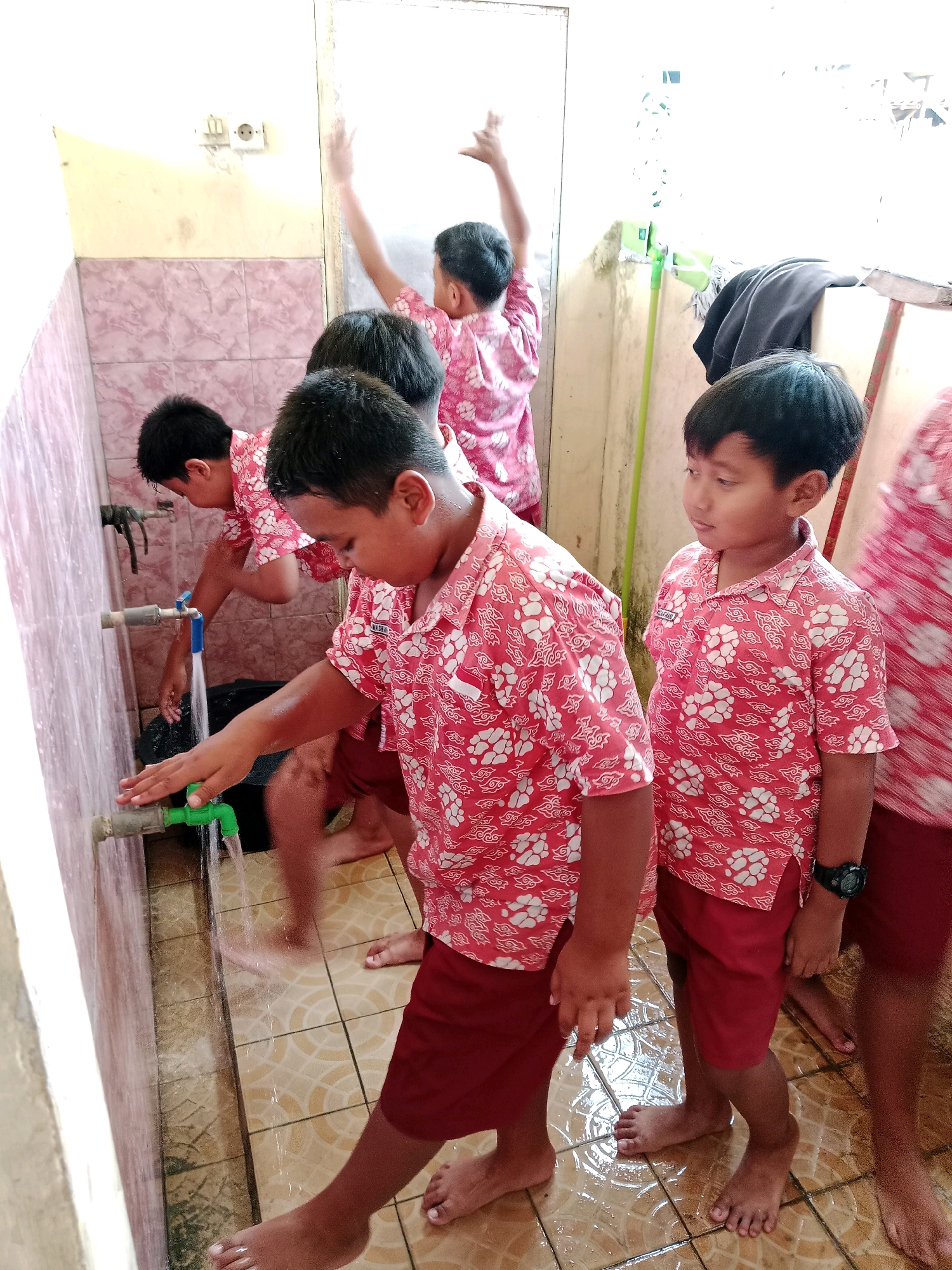 This image depicts a group of male children, likely students, engaged in washing their hands at a public faucet. They are wearing matching red shirts with a white cloud-like pattern and dark red shorts, suggesting a school uniform. The environment appears to be an outdoor or semi-outdoor area, possibly a restroom or wash area, with tiled flooring that is wet from the water. The walls are partially tiled with pinkish-brown tiles and partially plastered. In the background, a broom and dustpan are visible, along with a dark-colored garment hanging on a support. The children are interacting with the faucets, with one child in the foreground directly facing the camera, looking downwards at the water. The scene is likely located in Salatiga, Indonesia, given the context of school children in a tropical climate.