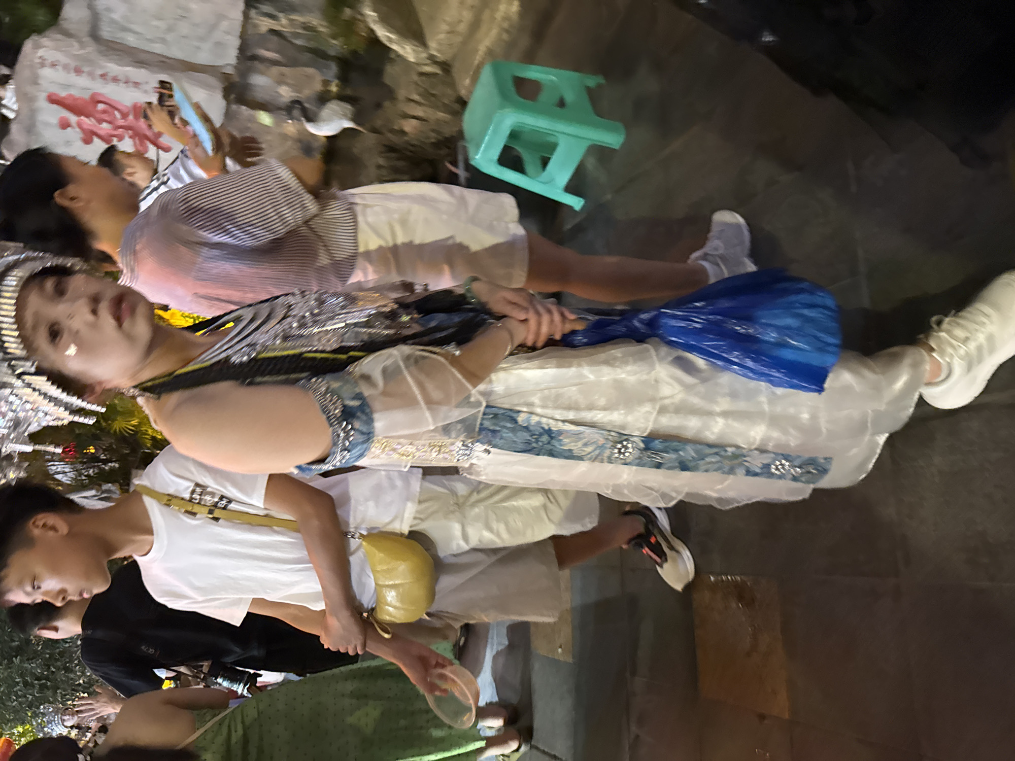 The image shows a group of people outdoors, possibly at a cultural event or market, with a focus on a young woman in elaborate attire. The woman in the foreground is wearing a white, layered dress with blue accents and a detailed headdress. She is holding a blue plastic bag in her right hand and her left arm is bent, with her hand resting on her hip. Beside her, a young boy in a white t-shirt and light-colored shorts is visible, carrying a yellow purse across his chest. Another woman in a striped shirt and white shorts is standing to the right of the boy, holding a phone. In the background, a green plastic stool is placed on the ground. The ground appears to be paved. There is a stone wall with red Chinese characters behind the people. The scene suggests a festive or cultural gathering in an urban setting, likely in the Yuzhong District of China, given the presence of Chinese characters and the urban environment.