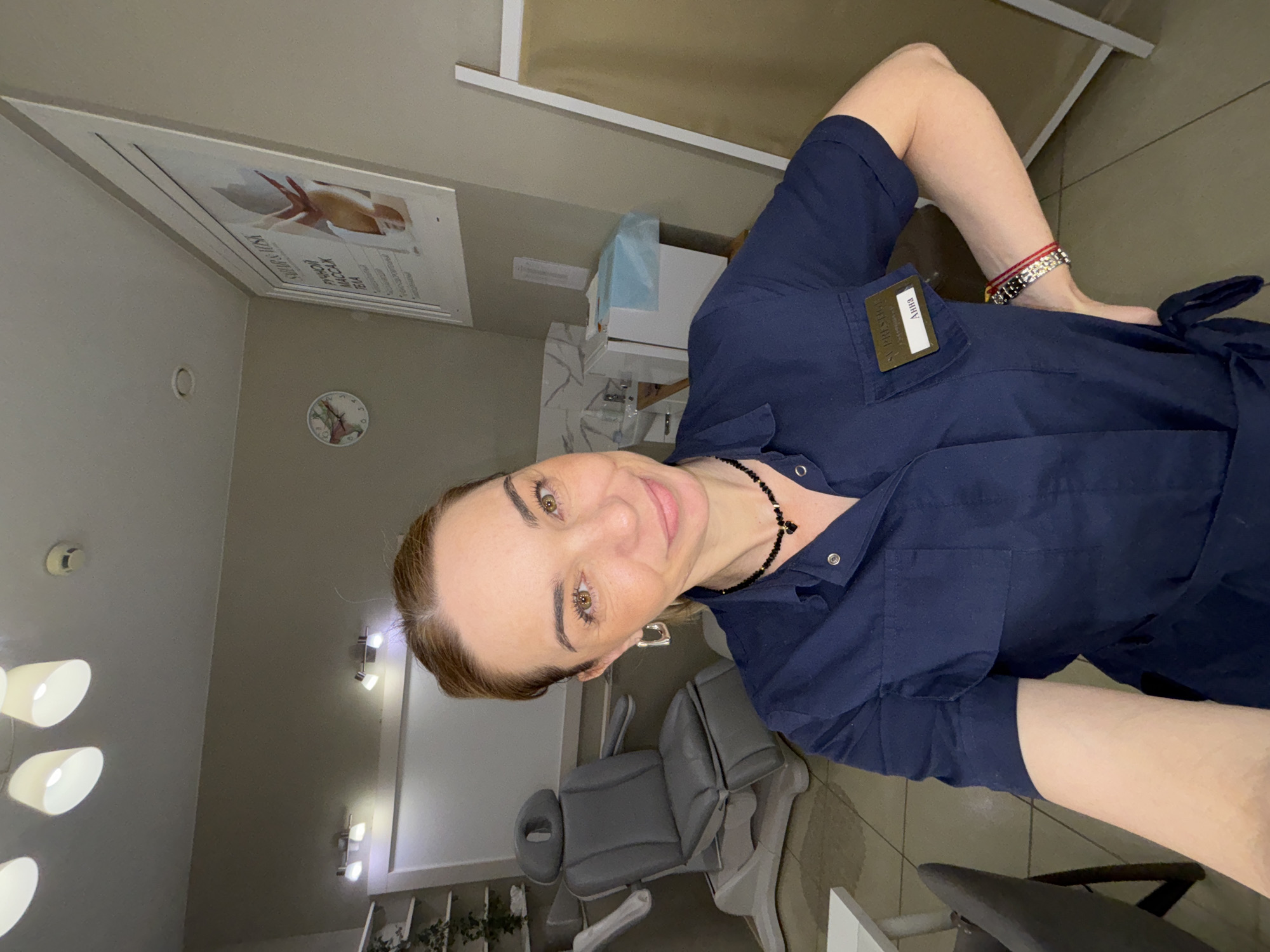 A woman takes a selfie in what appears to be a beauty salon or aesthetic clinic. The image is oriented sideways, with the woman positioned vertically on the left. She has light brown hair, styled back, and a gentle smile directed at the camera. Her eyes are brown. She wears a dark blue uniform or scrubs with a collar and snap buttons. A black beaded necklace with a central pendant is visible around her neck. On her right chest (left side from the viewer's perspective), she wears a rectangular name tag that clearly reads "АННА" (ANNA). Above her name, part of an English phrase like "SKIN EXPERT" is faintly visible, and below it, text likely indicating her profession, possibly "ЭСТЕТИСТ" (ESTHETICIAN). Her left arm is visible, adorned with multiple bracelets, including one with red and white beads and another metallic, possibly silver, design.
The setting is an indoor room with light-colored walls and tiled flooring. In the background, a grey upholstered treatment chair or bed with adjustable sections is visible, suggesting a professional setting for client services. A white cabinet or counter is also present, with a blue plastic bag, possibly containing supplies, on top. Lighting fixtures include a multi-bulb floor lamp with white cone-shaped shades on the left, and several wall-mounted spotlights on the right. A circular wall clock with a floral design is on the wall, along with a framed poster. The poster displays an image of a person's torso and visible Russian text, including parts of words like "МАССАЖ" (MASSAGE) and "РЕЗУЛЬТАТ" (RESULT), further supporting the beauty or wellness context. A ceiling-mounted device, possibly a smoke detector, is also visible. The woman's expression is pleasant and professional.