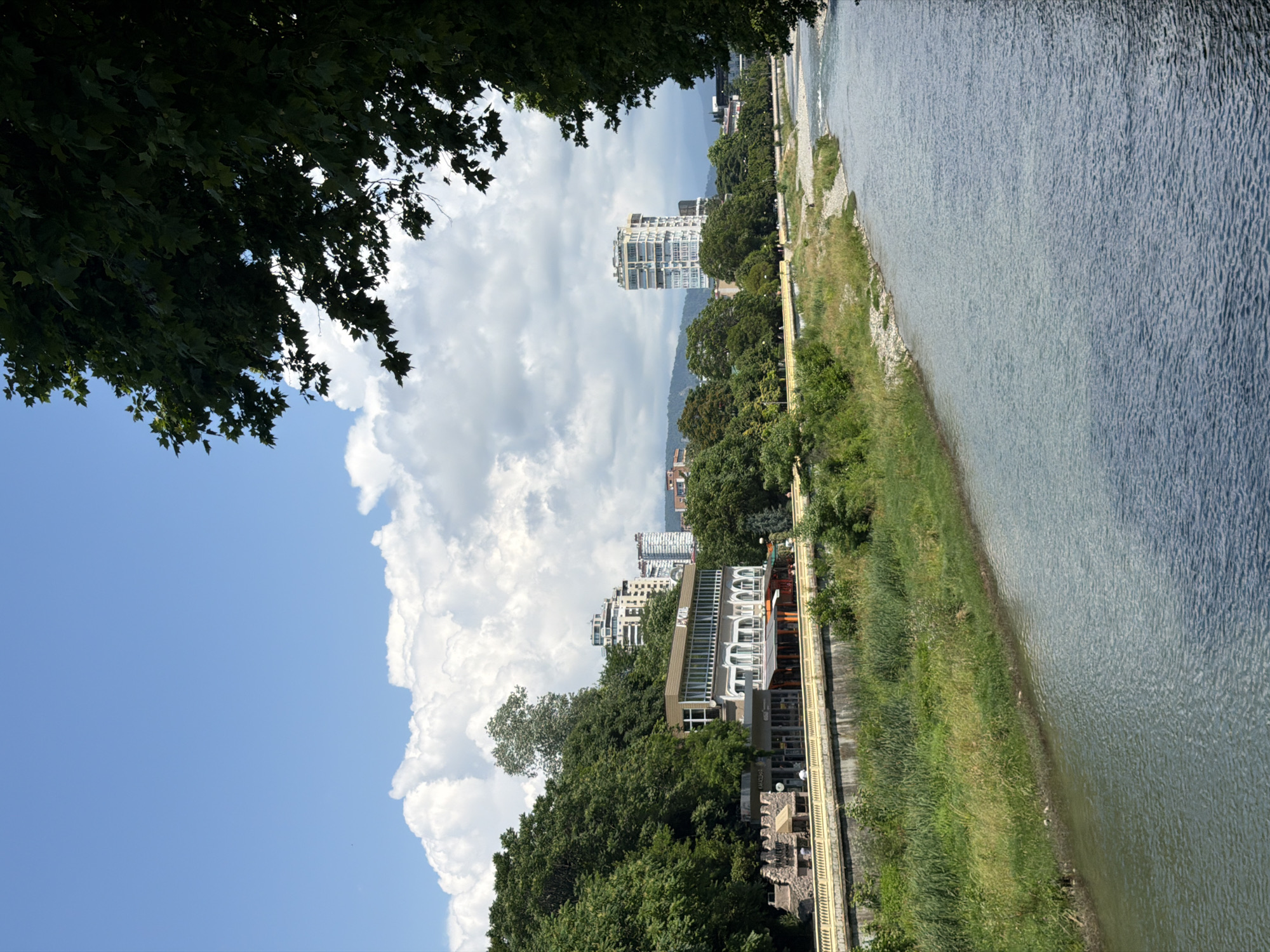 This is a vertical, eye-level shot of a scenic riverside promenade in Sochi, Russia. The image is dominated by lush green trees in the foreground, framing a view of a calm river on the right side. The river reflects the sky and surrounding landscape, with gentle ripples visible on its surface.
A paved walkway or promenade runs along the riverbank, bordered by a yellow railing. Several buildings are visible along this promenade, including a modern structure with a distinctive arched facade and a taller, white, multi-story building in the distance. The architecture suggests a developed urban area integrated with nature. The background features rolling hills and a hazy sky filled with large, fluffy white clouds, indicating fair weather.
The time of day appears to be daytime, likely mid-morning or afternoon, given the bright natural light and the shadows cast by the trees and buildings. There are no visible people in the frame, suggesting a relatively quiet moment. The overall atmosphere is peaceful and serene, highlighting the natural beauty of the location.
No discernible text is visible in the image. The scene captures a harmonious blend of urban development and natural scenery, characteristic of a well-maintained public space.
