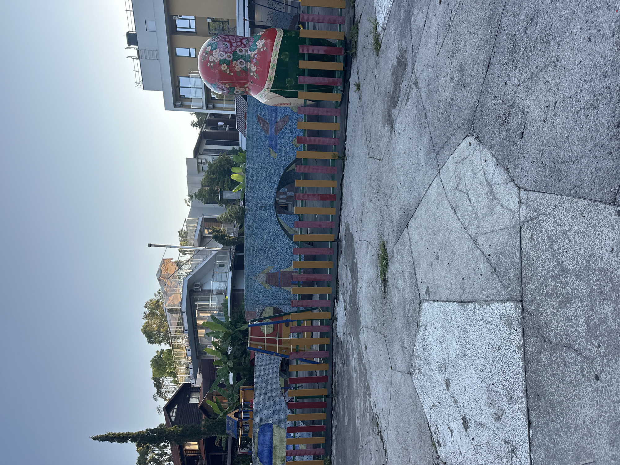 An outdoor shot captures a children's playground in Sochi, Russia, with a clear sky and lush green trees surrounding the area. The playground features a colorful fence and a large matryoshka doll, a traditional Russian nesting doll, standing prominently in the scene. The ground is paved with concrete, showing signs of age with cracks and worn patches. In the background, buildings of various architectural styles, including modern apartment blocks and houses, are visible. The overall atmosphere is one of a quiet, possibly early morning, scene in a residential area of Sochi.