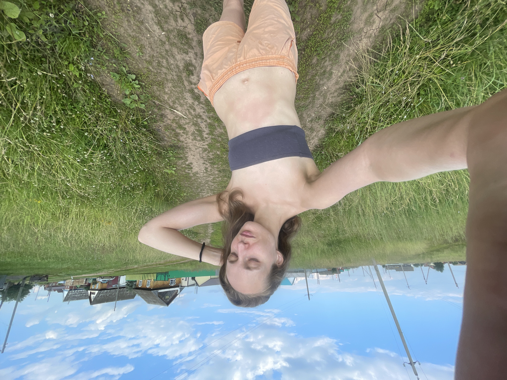 The image shows a young woman lying down in a grassy area with a dirt path. She is wearing peach-colored shorts and a dark gray bandeau top. Her eyes are closed, and her head is tilted back, with her hands resting behind her head. She appears relaxed and at peace. The background shows a clear blue sky with fluffy white clouds and a small village with wooden houses. The sun is shining, suggesting it is a warm and sunny day. The overall mood is serene and natural. The location is Kuyeda, Russia.
