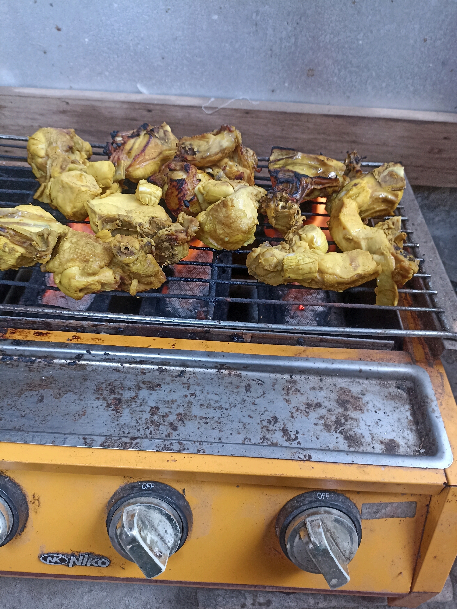 In Sonobekel, Indonesia, a close-up, high-angle shot captures chicken pieces being grilled over hot coals. The chicken, which appears to be marinated and seasoned with a yellow spice, is arranged on a metal grill rack. Visible beneath the rack are glowing red embers, indicating the heat source. The grill is placed on top of a yellow gas stove, which shows signs of wear and tear, with some rust and grease stains on its surface. The stove has three control knobs, two of which are clearly visible in the foreground. One knob is labeled "OFF." The background is a textured, light gray wall and a wooden plank. The scene is set outdoors, with the ground beneath the stove appearing to be concrete or pavement.