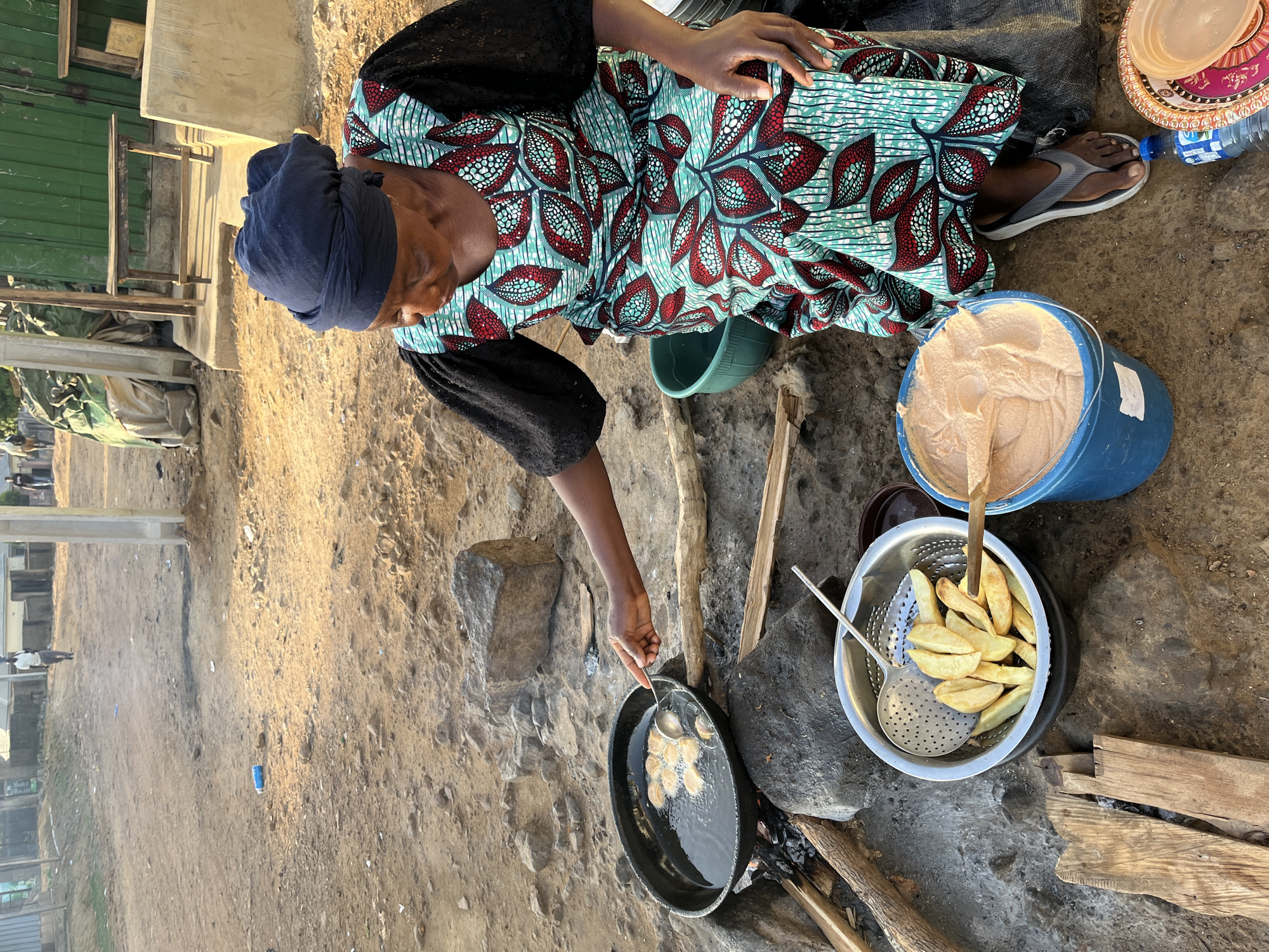 A woman wearing a dark blue headwrap and a teal, red, and black floral patterned dress is sitting on the ground in Jos, Nigeria, frying small dough balls in a large black pan over an open fire. To her right is a metal colander filled with fried potato wedges, a blue bucket filled with a light-colored paste, and a stack of plastic plates. She is using a spoon to turn the dough balls in the pan of oil. The ground around her is dirt and sand.
