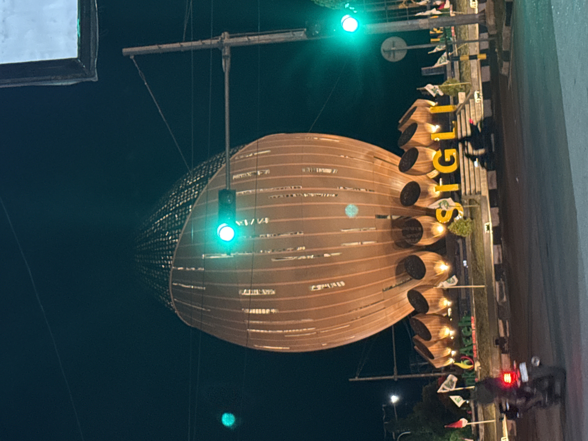 The image shows a nighttime view of the city of Sigli, Indonesia. The primary subject is a large, ellipsoidal structure, possibly a monument or building, constructed with horizontal bands and having multiple cylindrical protrusions on one side. The word "SIGLI" is spelled out vertically in large, illuminated yellow letters adjacent to the structure. Traffic lights are visible, with at least two showing green. A motorcycle with its rear light illuminated is on the road. Flags and decorative elements are also present.