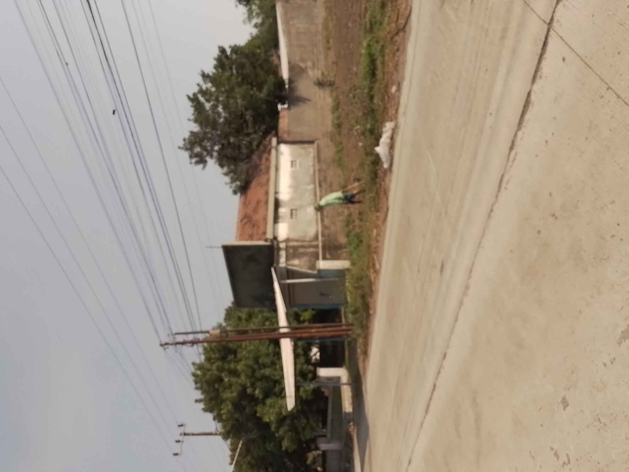 A person in a green shirt and dark shorts is using a rake on a patch of dry, brown earth near a low wall. Behind the person, a light-colored, single-story building with a reddish-brown tile roof is visible. A white plastic bag lies on the ground near the raking person. The scene is situated in a moderately populated area of Wates, Indonesia, with a paved road in the foreground and overhead power lines stretching across the sky. Mature trees with dense green foliage are present behind the building. The sky is a pale, hazy blue, suggesting a bright but possibly dusty day.