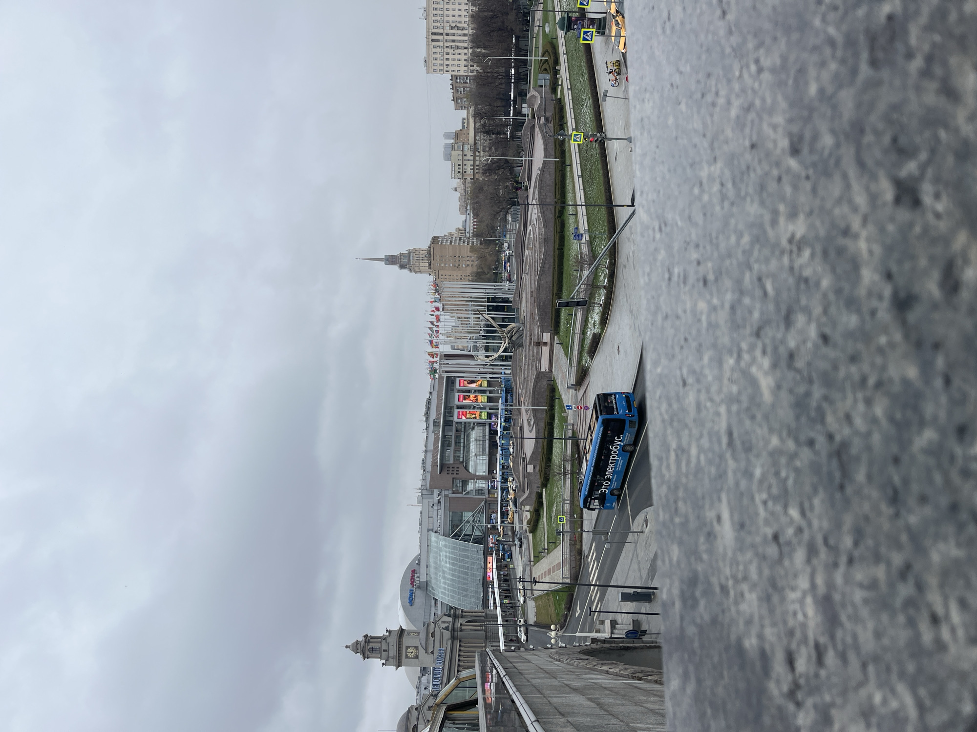 The image shows a cityscape, likely taken from a high vantage point in Moscow, Russia. The weather appears overcast. In the foreground, a textured grey concrete or stone structure, possibly part of a bridge or building, is visible. The primary subject is the urban scene extending into the distance.
Key elements include a large, modern building with a partially glassed-in roof, possibly a shopping center or transit hub. Nearby is a square with flags of various nations on display. A blue double-decker bus is traveling down the street, with the text "ЭТО ЭЛЕКТРОБУС" (This is an electric bus) visible on its side. Streetlights and traffic signals are positioned along the roads. A clock tower is visible, presumably marking the station. Several pedestrians and vehicles can be spotted in the background. Further away, various buildings form the skyline.