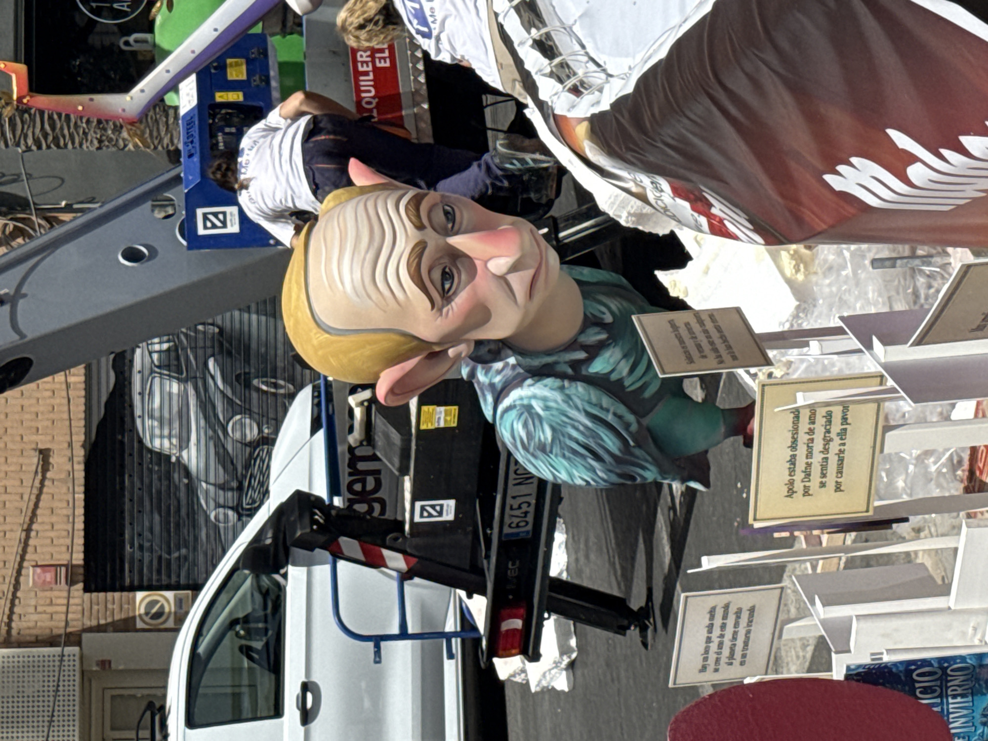 In an outdoor urban setting, a caricature of a bald man with large ears and wrinkled skin, resembling Vladimir Putin, is the central subject. The figure is depicted as a puppet or effigy, with a teal, feathery chest and red feet. It is positioned in front of a white vehicle and a mechanical lift. Several signs with text in a foreign language are visible around the effigy. In the background, a building with a rolling metal door features a mural of a vintage car. Two individuals, one wearing a white t-shirt and dark pants, and another partially visible woman with blonde hair, are near the mechanical lift. The ground is paved, and some debris is scattered around. The lighting suggests a daytime scene, likely in warm weather.