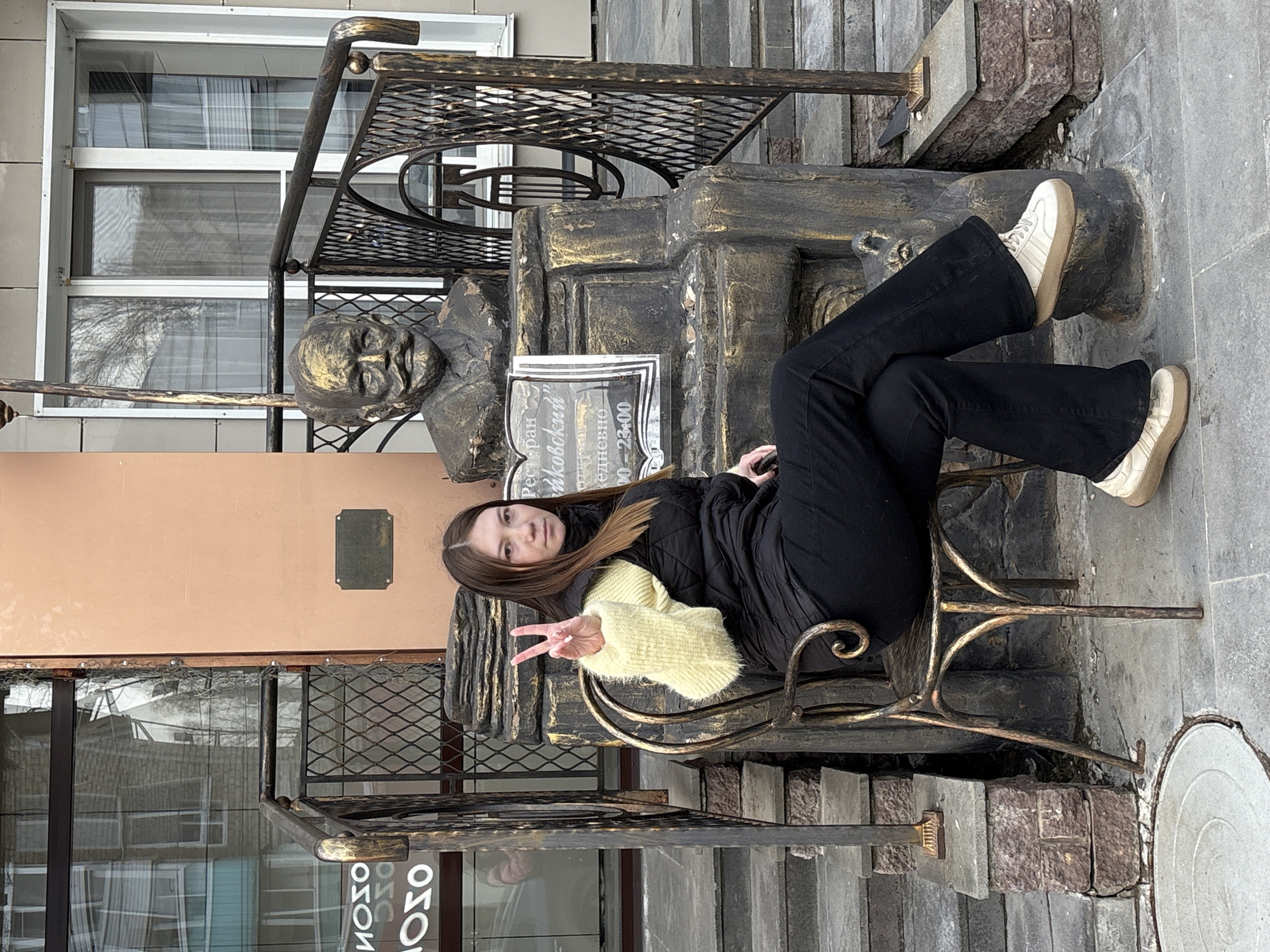 A young woman sits on a decorative metal chair next to a bust of Peter Ilyich Tchaikovsky, giving a peace sign. The chair is ornate and metallic, placed near a monument dedicated to the composer. The monument is on a small elevated platform, with steps leading up to it, and is surrounded by a metal railing. The background includes a building with windows and a peach-colored exterior wall. The location is in the city of Chaikovsky, Russia, as indicated by the subject of the monument.