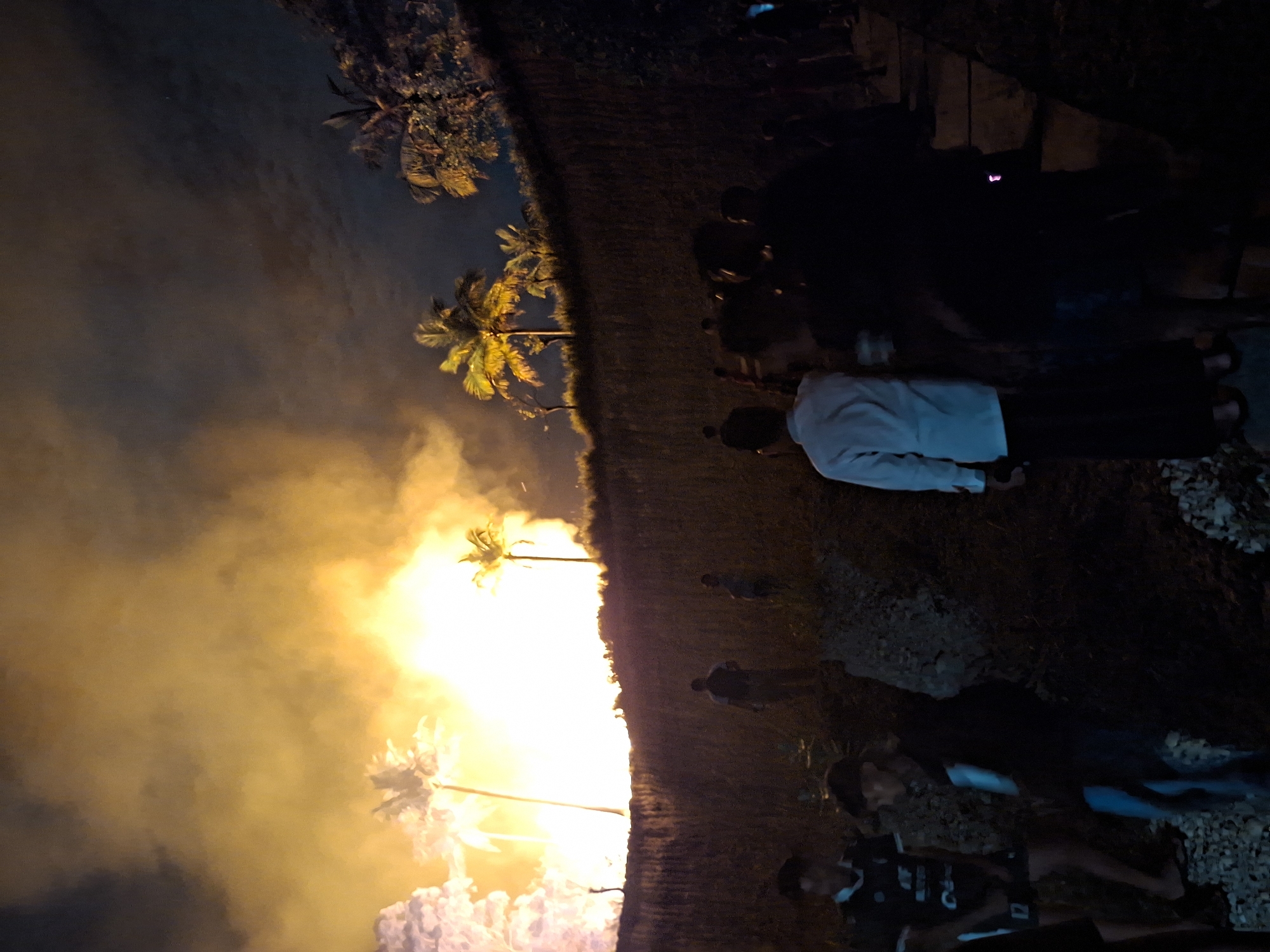 The image depicts a nighttime scene in Lhokseumawe, Indonesia, likely a bonfire event. A large fire is burning, illuminated with an orange glow, surrounded by smoke. Palm trees are visible near the fire. Several people are present, standing or observing. The ground appears to be a mix of dirt and gravel. Some artificial structures, possibly part of a building or platform, are visible in the upper right.