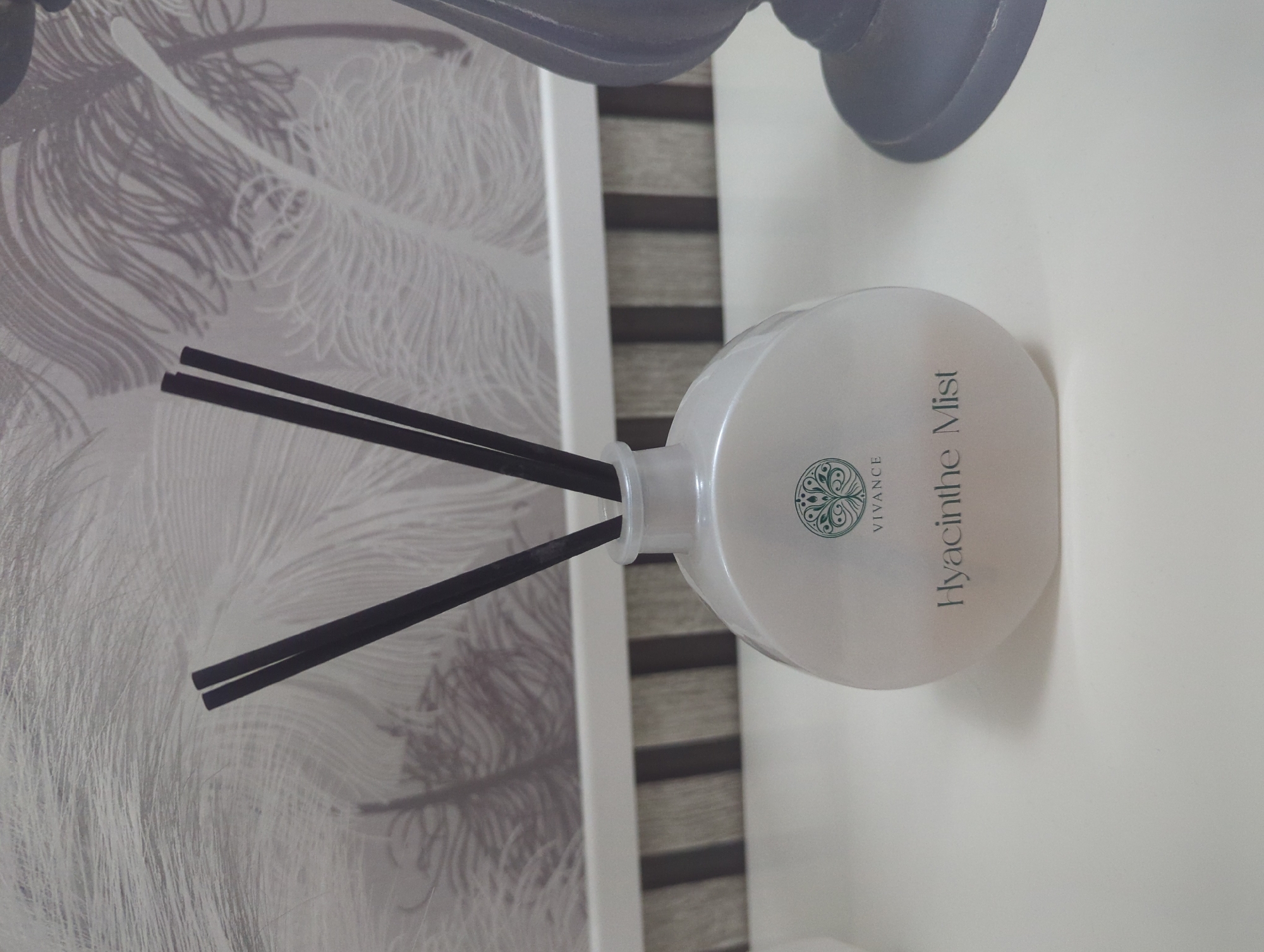 The image shows a white ceramic diffuser bottle with black reeds. The bottle has a circular green logo with the word "VIVANCE" above it, and "Hyacinthe Mist" written below. The diffuser is placed on a white surface, with a blurred background that appears to be a wall with a feather pattern. To the right of the diffuser, a dark grey decorative object, possibly a lamp base, is partially visible. The scene is indoors. The location is not specified in the image.