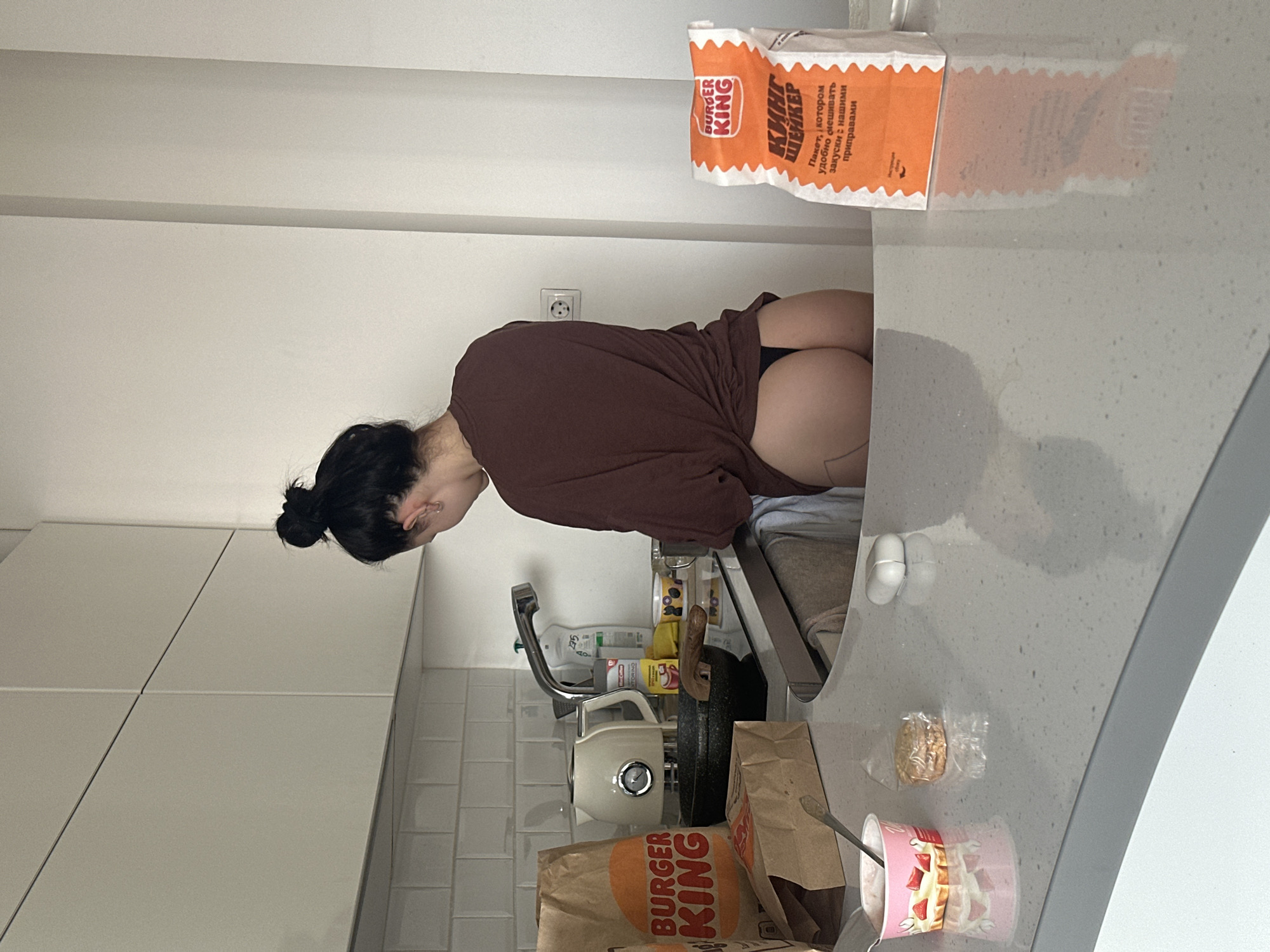 A woman with dark hair pulled into a bun is leaning over a kitchen sink. She is wearing a brown, loose-fitting top and dark underwear. The kitchen has white cabinets, a gray countertop, and tiled walls. A Burger King bag is visible on the counter near her back, and another Burger King bag is at the bottom left of the frame, along with a cup of strawberry yogurt and a wrapped pastry. A white electric kettle and various cleaning supplies are near the sink. Another Burger King bag is standing up against the wall. There is an electrical outlet on the wall. The location appears to be a kitchen in a home.