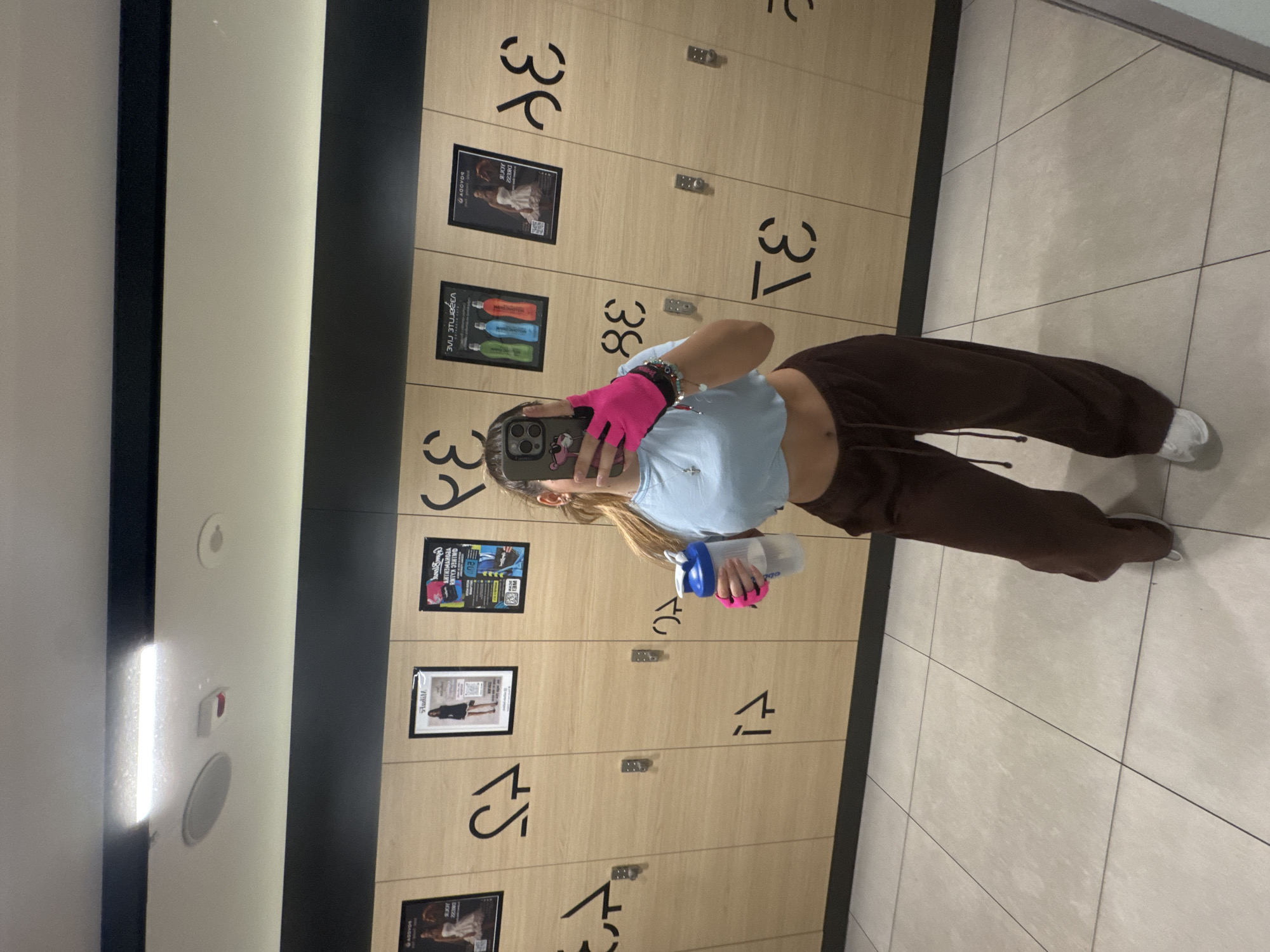 The image depicts a woman taking a mirror selfie inside a locker room at a Pulse Fitness & Spa facility in Sofia, Bulgaria. The subject, seen from the mid-torso down, has her face obscured by her smartphone, which she holds in her right hand. She is wearing a light blue crop top, dark brown wide-leg sweatpants with a drawstring, and white athletic shoes. A silver cross necklace is visible, along with multiple bracelets on her right wrist. Her left hand holds a white and blue shaker bottle, and she wears pink fingerless workout gloves on both hands. Her smartphone has a pink panther sticker on the back.
The environment consists of light wood-paneled lockers with distinctive black stylized numbers and symbols (e.g., '75', '74', '38', '37', '36', '35') arranged in rows, indicating individual storage units. Each locker has a small silver handle/lock. Several framed posters are affixed to the lockers; due to the mirror reflection, their text appears reversed. Visible elements include a poster with a model in a white dress (text 'ASSOVCA' partially visible), another with a model in a black dress (text 'Andil' partially visible), and a colorful poster showing bottles, likely for supplements (reversed text '3VJI 3TJUG3A' visible). The floor is light grey tiled with dark grout lines. The ceiling, partially visible in the reflection, is white and features a long rectangular LED light fixture, smaller recessed circular lights, and a red sensor or indicator.