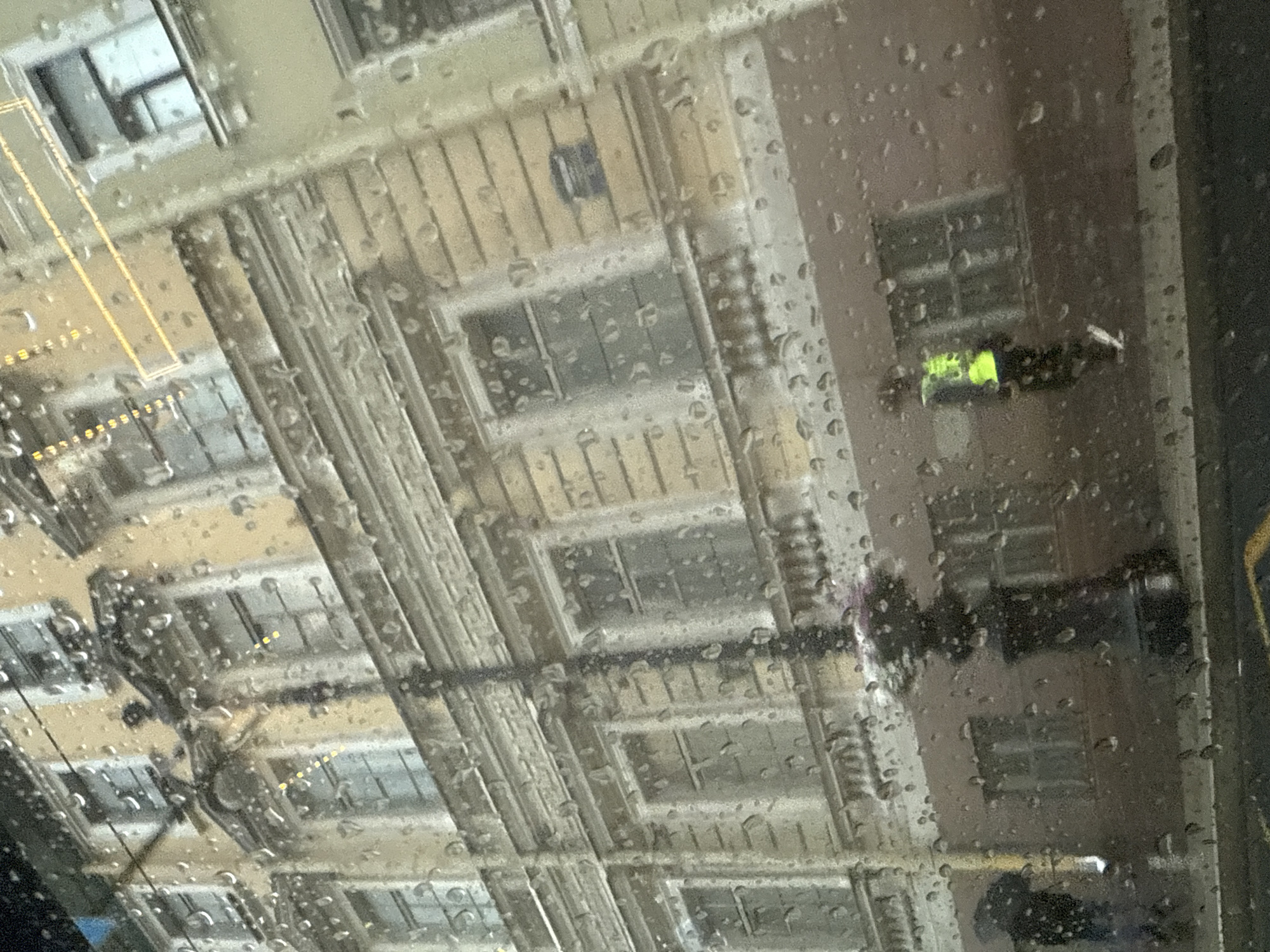The image depicts a view through a rain-streaked window onto a building facade in Saint Petersburg, Russia. The architecture suggests a historic building with multiple stories, ornate trim, and numerous windows. The windows themselves appear to be multi-paned and are reflecting the overcast, rainy sky. In the foreground, water droplets distort the view. Visible on the street level are several figures. One individual, on the right side of the frame, is wearing a bright yellow-green reflective vest and dark pants, suggesting they might be a construction worker or someone involved in road maintenance. Another figure, on the left, is partially obscured and appears to be holding an umbrella. A lamppost with an intricate design stands prominently in the middle ground. The overall impression is one of a typical urban scene in Saint Petersburg during inclement weather.