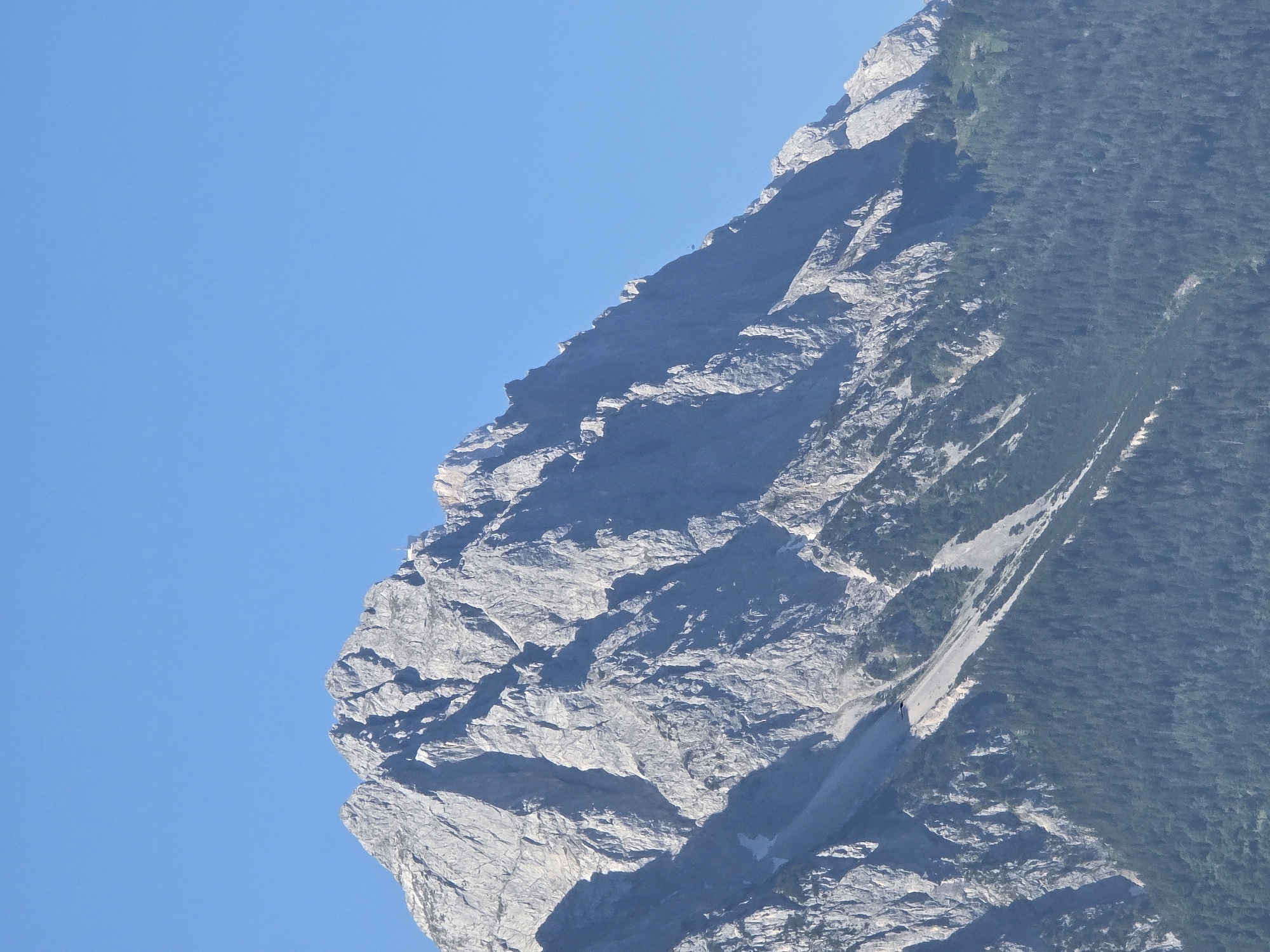 The image shows a close-up view of the Zugspitze mountain, the highest peak in Germany, located near Garmisch-Partenkirchen. The mountain's rocky, steep slopes are visible, with sparse vegetation lower down. The sky is clear and blue. There is a small building or structure at the summit and a cable car visible in the distance, indicating human presence and infrastructure.