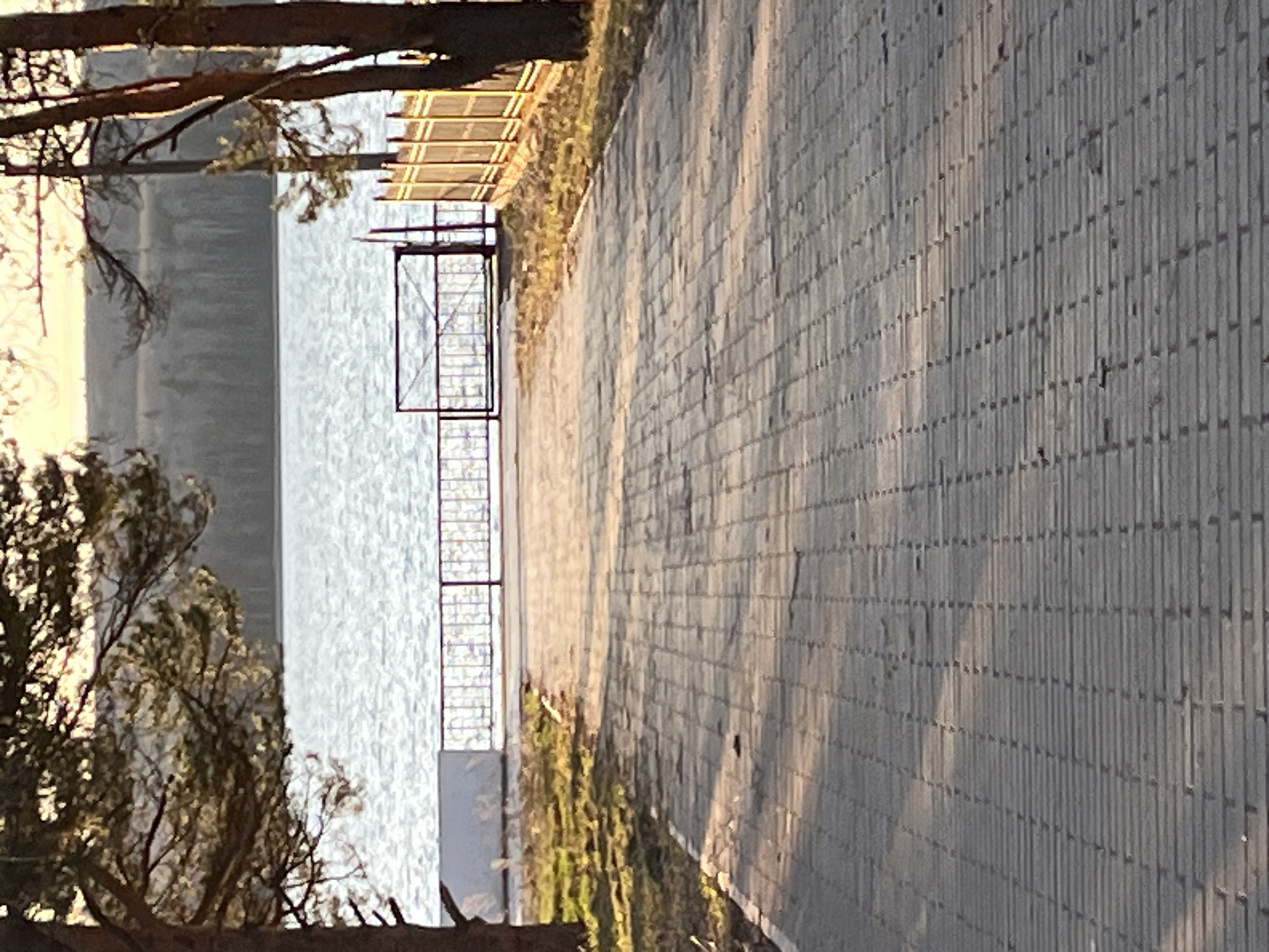 This image captures a tranquil outdoor scene, likely a riverside or lakeside path in Chaikovsky, Russia. The dominant feature is a long paved pathway, constructed from rectangular blocks, which extends from the right foreground towards the background. Long shadows are cast across the path, indicating either late afternoon or early morning sunlight. To the left of the path, a calm body of water, possibly a river or lake, reflects the warm, bright light, showing gentle ripples on its surface.
A dark metal structure, resembling a railing or a small pier, extends from the edge of the paved path towards or over the water. Branches and foliage from trees are visible in the top and bottom sections of the frame, framing the scene. The lighting suggests a clear, sunny day. There are no people visible, and no discernible text is present. The overall atmosphere conveyed is one of peace and natural serenity.
