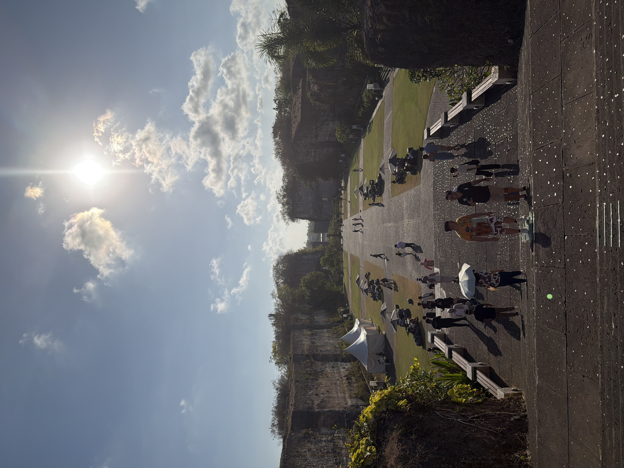 The image depicts a wide, paved staircase leading down to a plaza area with grassy sections, flanked by steep, rocky cliffs on either side. The sun is shining brightly in a blue sky with scattered clouds, casting long shadows. Several people are visible on the stairs and in the plaza.
In the foreground, on the stairs, a group of people are walking down. Two men are in the lead, one wearing a yellow t-shirt with a graphic print and patterned shorts, the other in a dark polo shirt and khaki shorts. Behind them, a person with an umbrella is walking, and further down, more individuals are seen ascending and descending.
The plaza area below features several white tents and what appear to be seating arrangements with umbrellas on the grass. There are also benches along the paved areas. More people are scattered throughout the plaza, some walking, others seated. The cliffs on both sides are covered in dense vegetation.
The overall impression is a scenic, open-air venue, likely a tourist attraction or park, given the presence of visitors and event-style tents. Based on the description, this location could be the Uluwatu Temple area or a similar cliffside attraction in Kuta Selatan, Indonesia, known for its dramatic coastal landscapes.