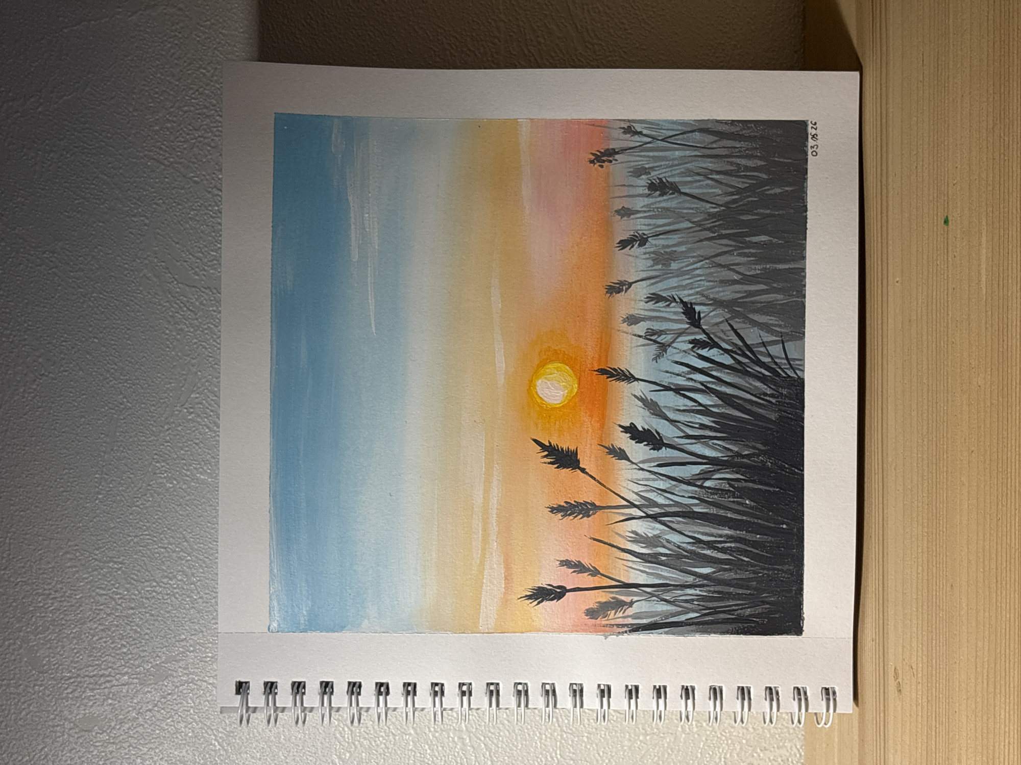 The image features a vibrant painting, likely watercolor or gouache, displayed on a page from a spiral-bound sketchbook. The painting depicts a serene landscape scene, capturing either a sunrise or sunset. The sky transitions from a soft blue on the left to warm shades of yellow, orange, and a touch of pink on the right. A luminous, circular sun, depicted in bright yellow with a white highlight, is positioned towards the lower right quadrant of the sky. In the foreground, tall, dark silhouetted grass or reeds with some lighter grey detailing are prominently featured, creating a strong contrast against the colorful sky.
The painting is presented within the white border of a sketchbook page, which is open. The white plastic spiral binding of the sketchbook is visible along the bottom edge of the image. To the left of the sketchbook, a textured, light grey surface is visible, while a vertically grained wooden surface appears on the right. There are no people present in the image. In the top right corner of the painting's white border, the date "03.05.20" is written vertically. The overall mood conveyed by the painting is tranquil and atmospheric. While the provided context indicates the location is Perm, Russia, no specific geographical cues are visible within the image itself.