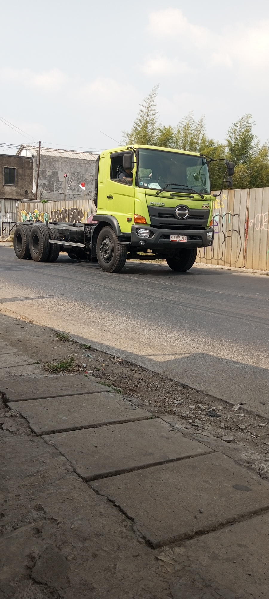 A bright yellow Hino 200 truck, a chassis cab model, is driving on a paved road in Setu, Indonesia. The truck is on the right side of the road, moving towards the viewer. The driver is visible inside the cab. The truck has multiple axles with dual rear wheels. The background includes a concrete barrier with graffiti, some overgrown vegetation, and a building with a window. The sky is partly cloudy. The foreground shows a cracked concrete sidewalk with some weeds and debris.