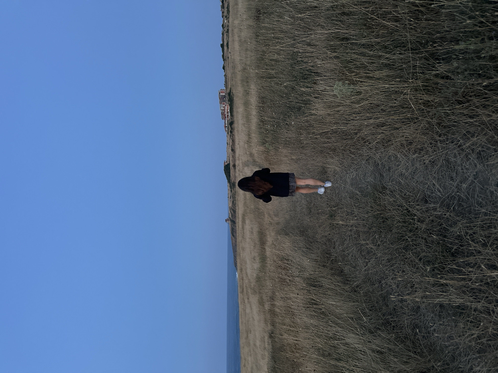 A person with long, dark hair is standing in a field of dry, tall grass, facing away from the camera. They are wearing a dark hooded jacket and a skirt, and white sneakers. The field stretches towards a body of water, which appears to be the sea, with a clear blue sky above. In the distance, along the coastline, there are buildings that suggest a populated area, possibly a resort town. A small tower or structure is visible on a hill further down the coast. The lighting suggests it is late afternoon or early evening, as the light is soft and the colors are muted. The overall atmosphere is peaceful and serene. The location is identified as Sinemorets, Bulgaria. There is no visible text in the image.