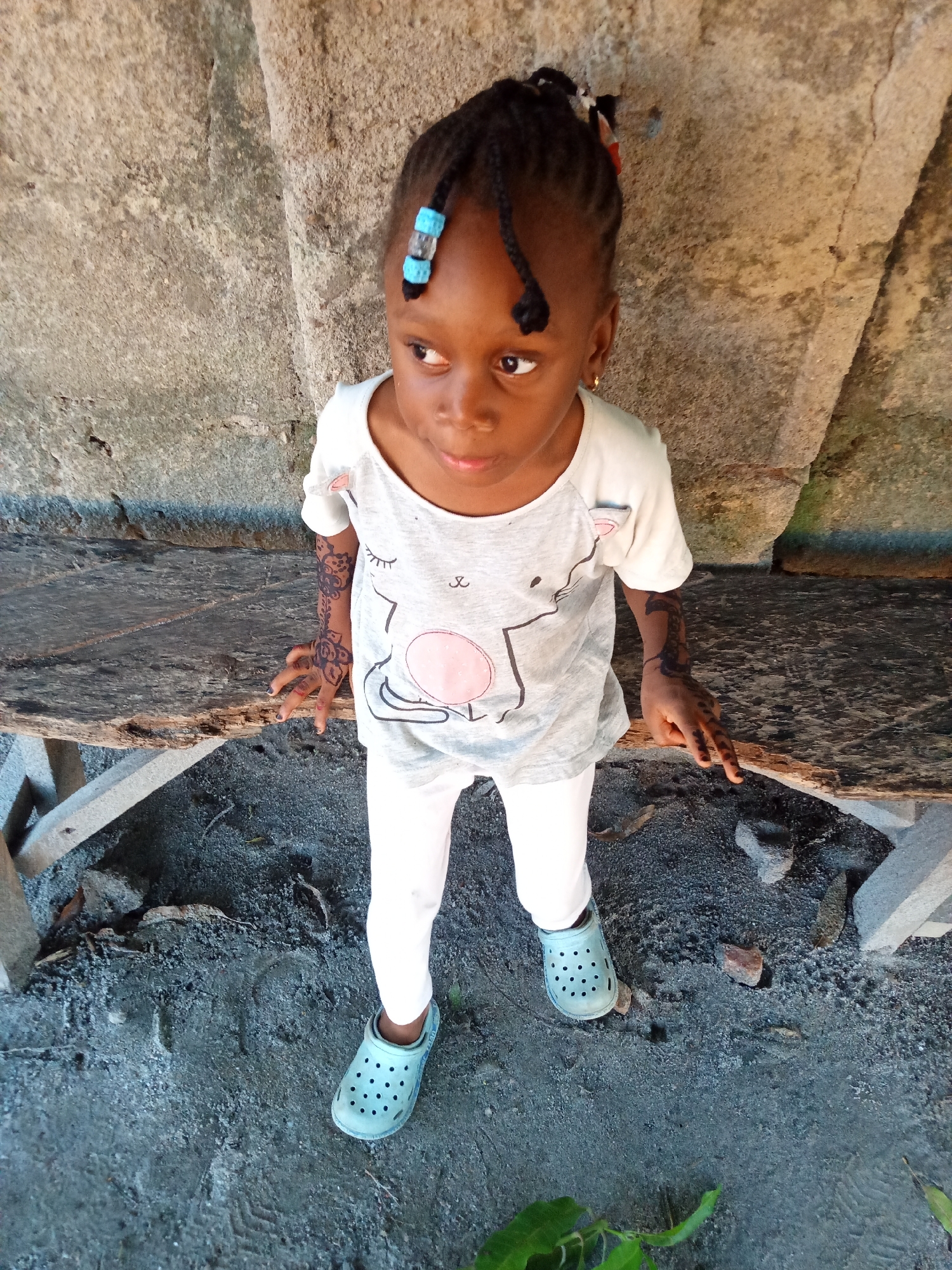 A young girl stands in front of a rough-hewn bench, likely in an outdoor location in Unknown, Unknown. She wears a gray t-shirt with a cat graphic, white leggings, and light blue Crocs. Her hair is styled in braids, some adorned with blue beads. There are decorative designs on her hands and lower arms. The bench sits on a surface covered in dark, coarse material, possibly sand or gravel. Behind the bench is a textured, weathered wall.