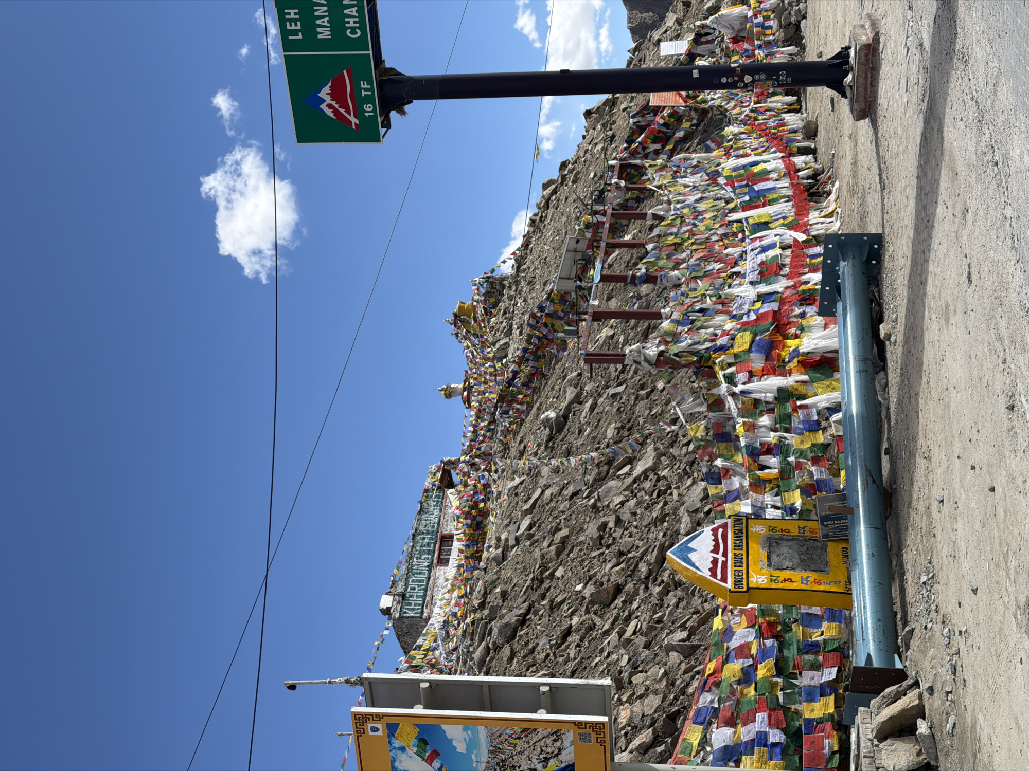 The image depicts a high-altitude mountain pass in Leh, India, characterized by a rugged, rocky terrain under a bright blue sky with scattered white clouds. The scene is dominated by prayer flags in various colors—red, blue, yellow, green, and white—strung across the mountainside and draped along fences, creating a vibrant tapestry against the muted rock and sky.
Prominently featured is a signpost with a green rectangular top, bearing a red and white mountain graphic and text that appears to read "LEH MANA CHANG 16 TF". To the left, another sign displays a colorful image of prayer flags and mountains, alongside a QR code and the text "KHARDOING LA". A yellow, triangular structure with the inscription "BORDER ROADS ORGANISATION" is also visible, adorned with Tibetan script.
In the foreground, a rough, unpaved road or path leads towards the mountain. A light blue metal barrier, likely a guardrail, runs horizontally across the lower part of the image. The overall atmosphere suggests a remote, spiritual, and windswept location, typical of mountain passes in the Himalayas. No people are visible in the frame.