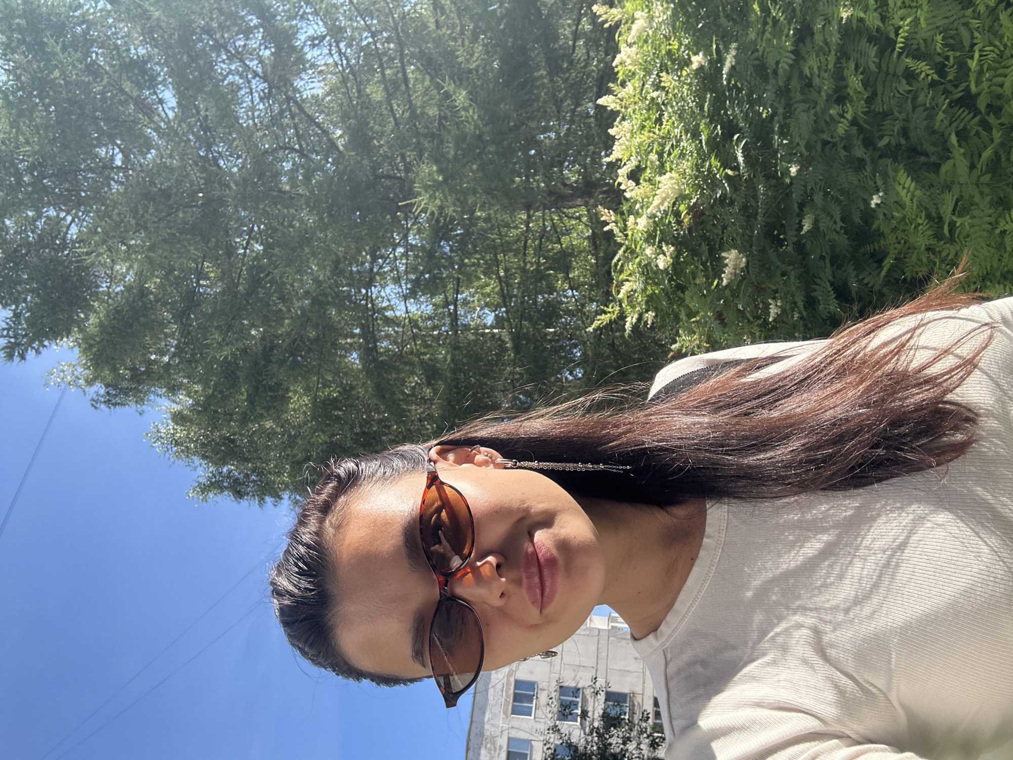The image is a selfie taken outdoors in Chaikovsky, Russia, on a sunny day. A young woman with long, dark brown hair is in the foreground, wearing a white long-sleeved shirt and tortoise-shell sunglasses. She has a dangling silver earring in her right ear. Behind her, a large evergreen tree dominates the background, with a building and a flowering bush visible to the left and right respectively. The sky is clear and blue, with some power lines visible across the top of the frame. The scene appears to be in a residential area, possibly a park or a yard, given the presence of the building, tree, and bush.