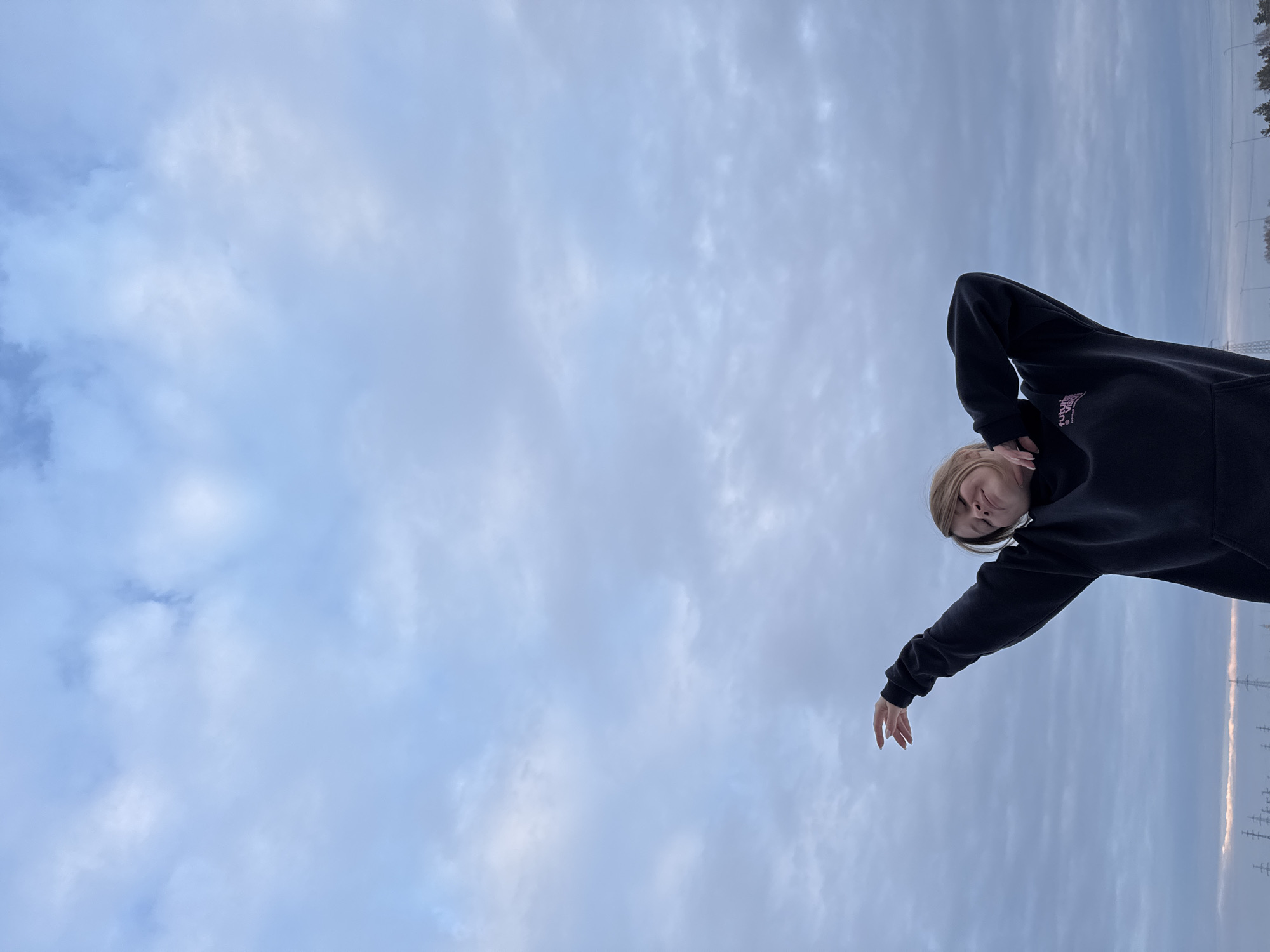 The image depicts a single individual in an open outdoor environment, captured from a low-angle perspective against an overcast sky. The specific location in Noviy, Russia, is stated in the prompt but not explicitly identifiable from visual cues within the image.
**Subject:** A fair-skinned female with blonde hair is positioned in the lower center of the frame. Her head is tilted back and slightly to her right, eyes closed or heavily squinted, with a slight upturned expression at the corners of her mouth. She is wearing a dark-colored hooded sweatshirt with a front pouch pocket. A partial light-colored logo, appearing to read "future vision," is visible on the left chest area of her garment.
**Actions and Pose:** Her left arm is bent at the elbow, with her left hand resting on the back of her neck. Her right arm is extended upwards and to her right, with the hand open and fingers slightly splayed, palm facing upwards. This posture suggests an act of stretching or a posed gesture.
**Environment:** The upper two-thirds of the image are dominated by a cloudy sky, featuring layered formations of blue-grey and white clouds, indicating overcast conditions. A subtle, lighter hue is present near the horizon on the far left. Along the distant horizon line at the very bottom of the frame, faint, dark silhouettes of multiple electricity transmission towers (power lines) are visible, interspersed with indistinct dark shapes suggestive of distant trees or structures. The ground is not visible. The overall lighting is diffuse.