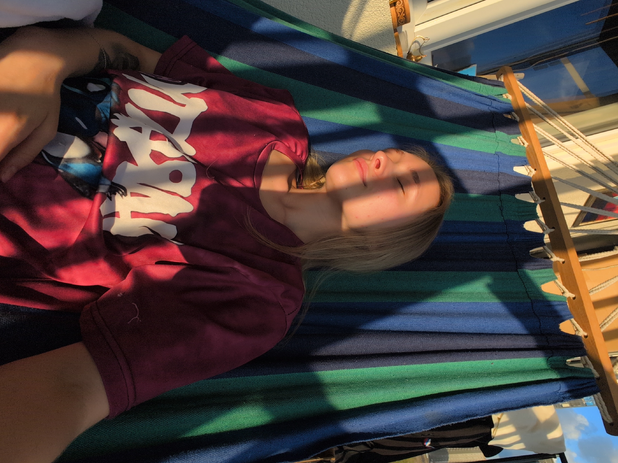 A young woman with blonde hair is lying in a hammock outdoors. She is wearing a maroon t-shirt with a white graphic print and has her eyes closed, appearing relaxed. The hammock is made of striped fabric in shades of blue and green. Sunlight casts diagonal shadows across her face and the hammock. In the background, there is a balcony railing, a window, and what appears to be a building. The overall scene suggests a peaceful, sunny day.